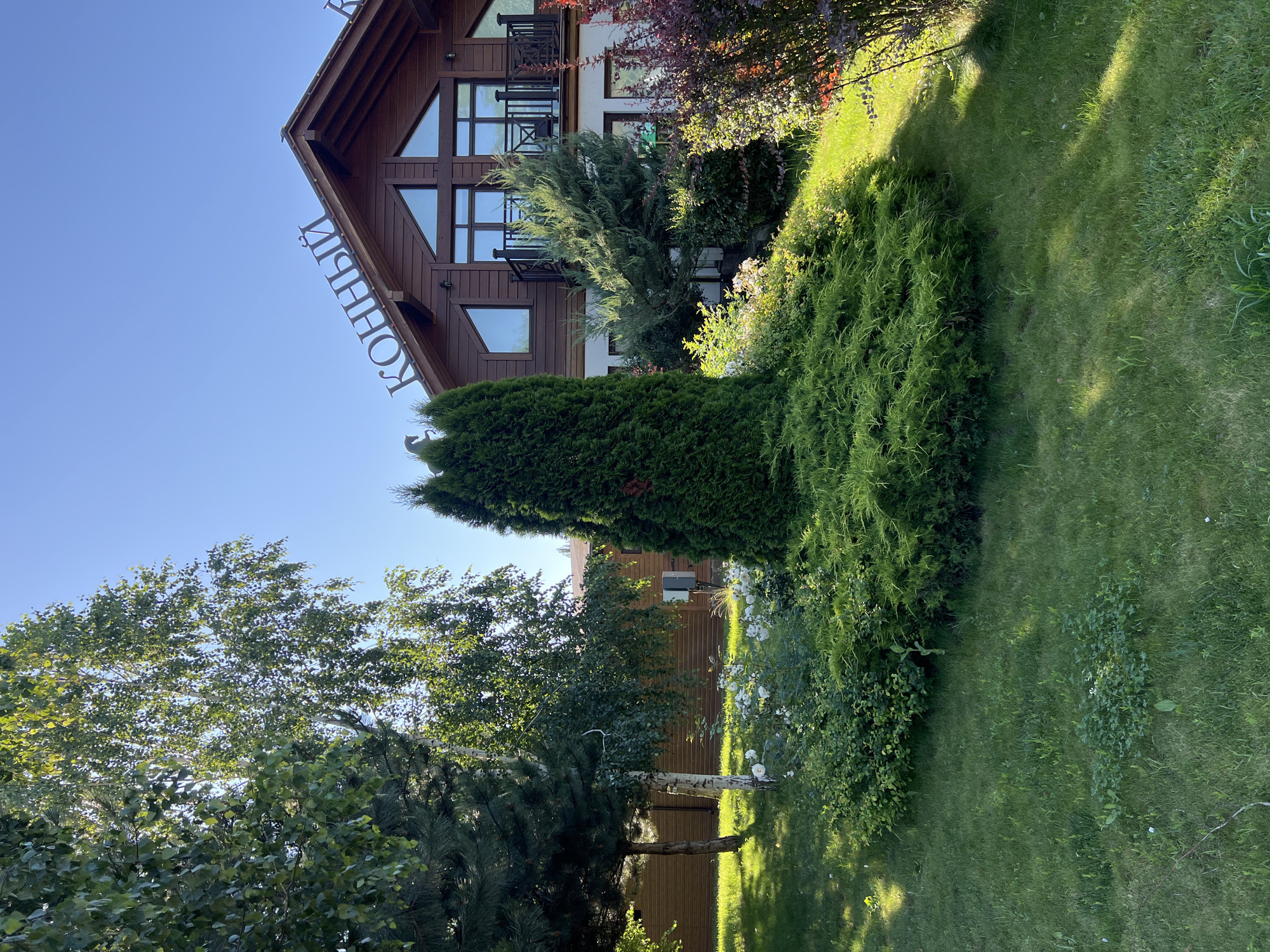 The image depicts the exterior of a wooden building, likely a commercial establishment given the prominent lettering on the roof that reads "КОННЫЙ" which translates to "Equestrian". The building has a rustic, wooden facade with large windows and balconies. In the foreground, there is a well-maintained garden with lush green grass, various shrubs, and a tall, conical evergreen tree. The scene is bathed in sunlight, casting shadows on the lawn, suggesting a clear, sunny day. The background shows more of the wooden building and a clear blue sky. The location is identified as Plastunivska, Russia. There are no people visible in the image.