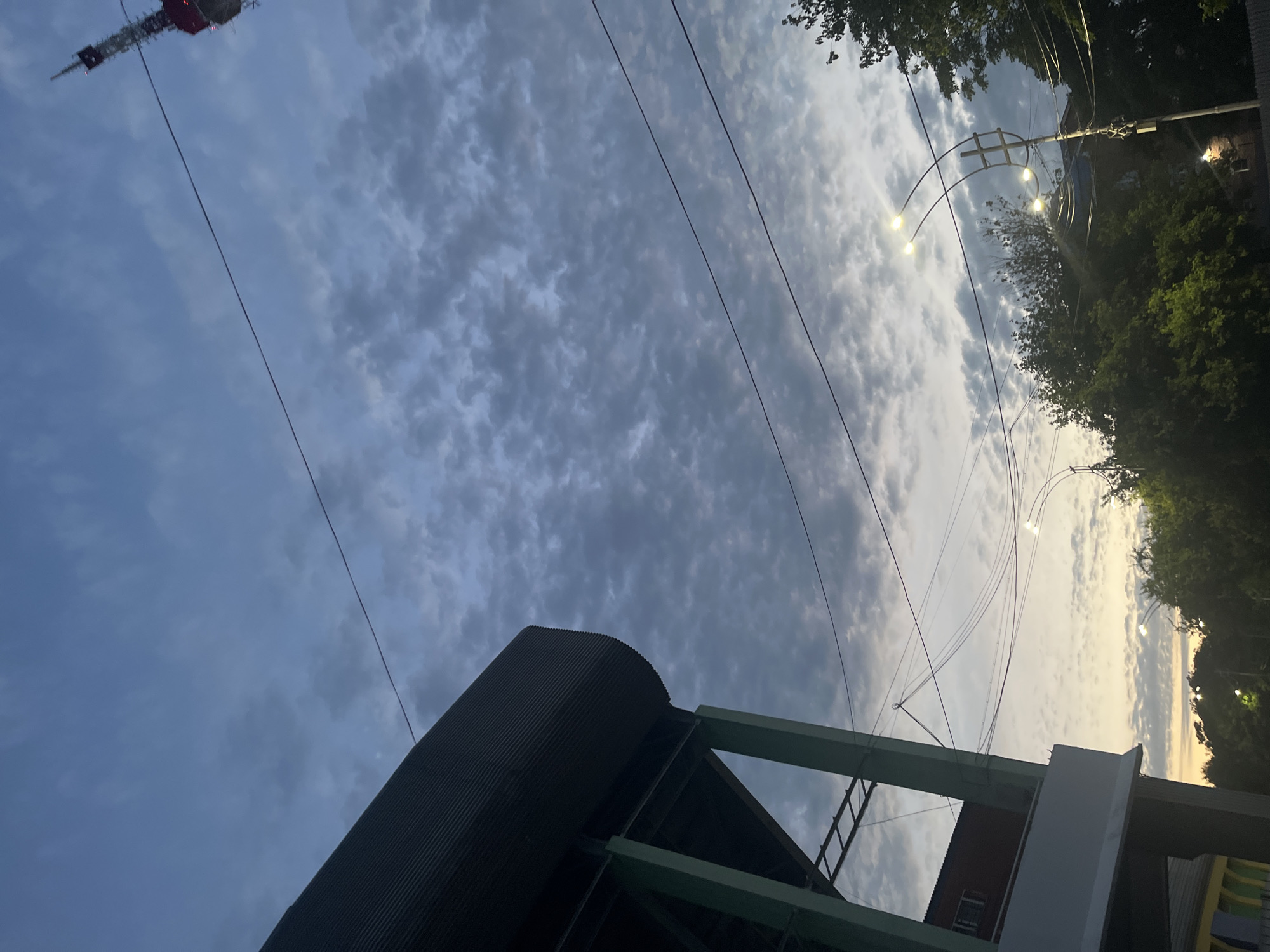 The image shows an outdoor scene, likely in Krasnodar, Russia, with a dramatic sky and a mix of infrastructure and natural elements. The sky is filled with textured clouds, suggesting it might be late afternoon or early evening, with the sun peeking through in some areas, creating a soft, diffused light.
In the foreground, there's a large, dark, cylindrical structure with a corrugated metal exterior, possibly a silo or a part of an industrial building. A green-painted metal framework, part of another structure, extends from the dark cylinder, with a ladder visible.
Overhead, several power lines stretch across the frame, some taut and others sagging slightly. Decorative outdoor lights, resembling multiple bulbs on curved arms, are also visible, adding an urban touch to the scene. Trees with lush green foliage frame the right side of the image, contrasting with the man-made elements. In the upper left corner, a tall transmission tower or antenna pokes into the sky, its red and white segments hinting at its scale.
The overall impression is one of urban landscape meeting the elements, with a sense of twilight approaching. No people are visible. There is no discernible text in the image.