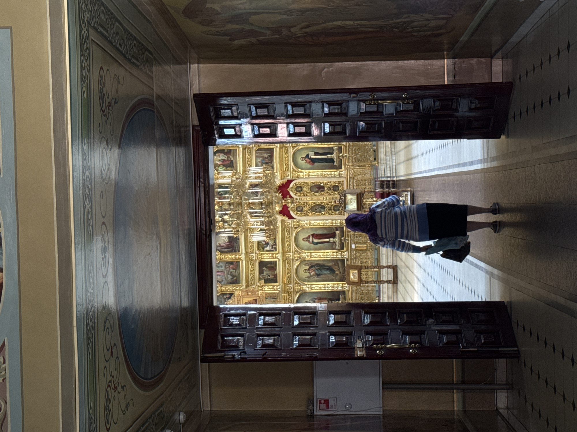 A person walks through a large wooden doorway into a church nave. The person, viewed from behind, wears a striped long-sleeved shirt and a dark skirt. They carry a dark tote bag in their right hand. The doorway opens into a brightly lit church interior featuring an iconostasis adorned with numerous golden icons. A chandelier hangs above the iconostasis. The church walls are decorated with religious paintings, and the ceiling is painted with celestial imagery. The floor of the church is tiled. The scene is captured from a low angle.