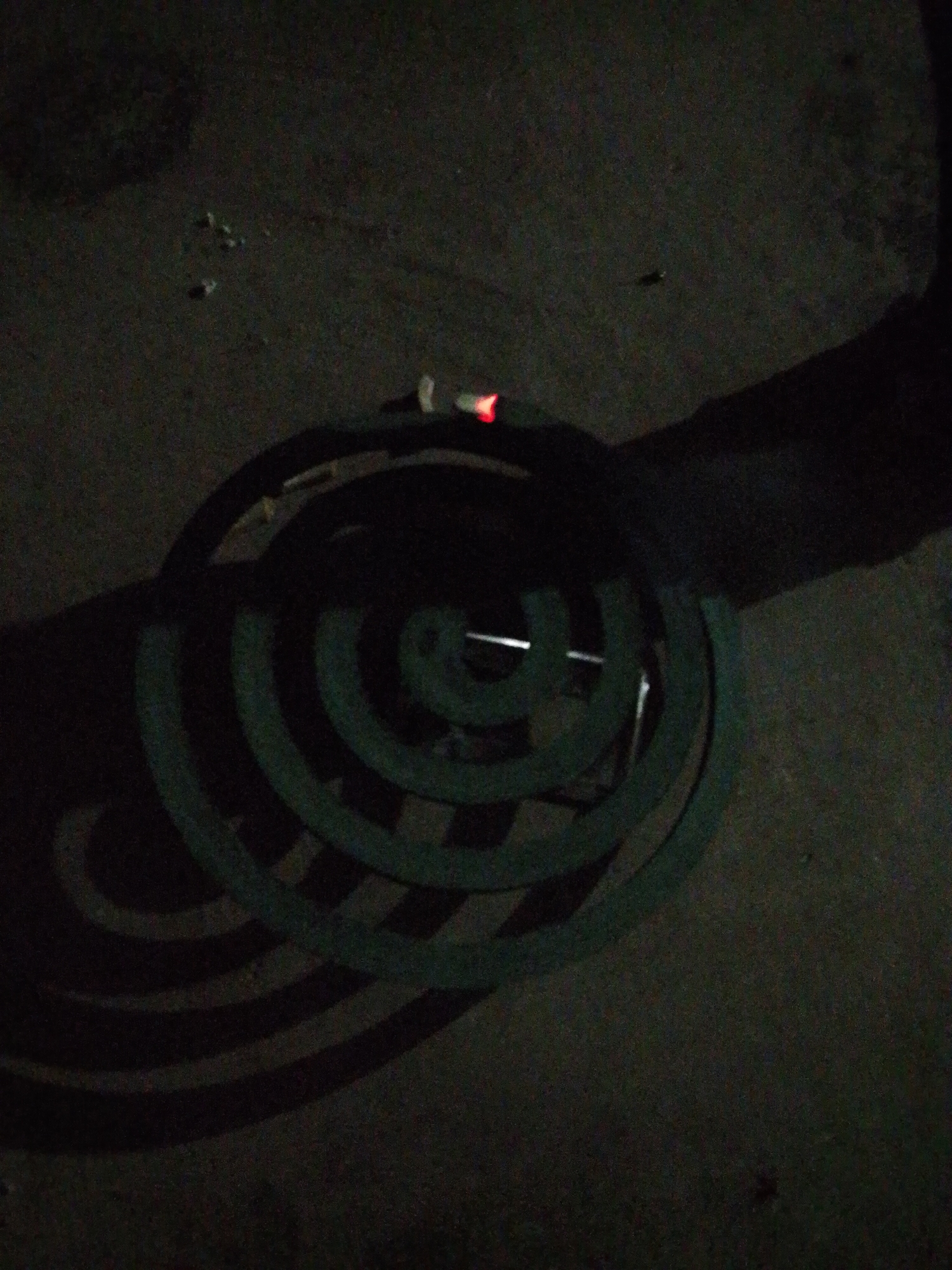 A close-up shot of a burning mosquito coil is placed on an outdoor surface at night, likely in Bukit Ketok, Indonesia. The dark environment is dimly lit, highlighting the red ember at the tip of the coil, which is supported by a metal stand. The coil's concentric circles cast distinct shadows on the ground. The surface appears to be rough and uneven.