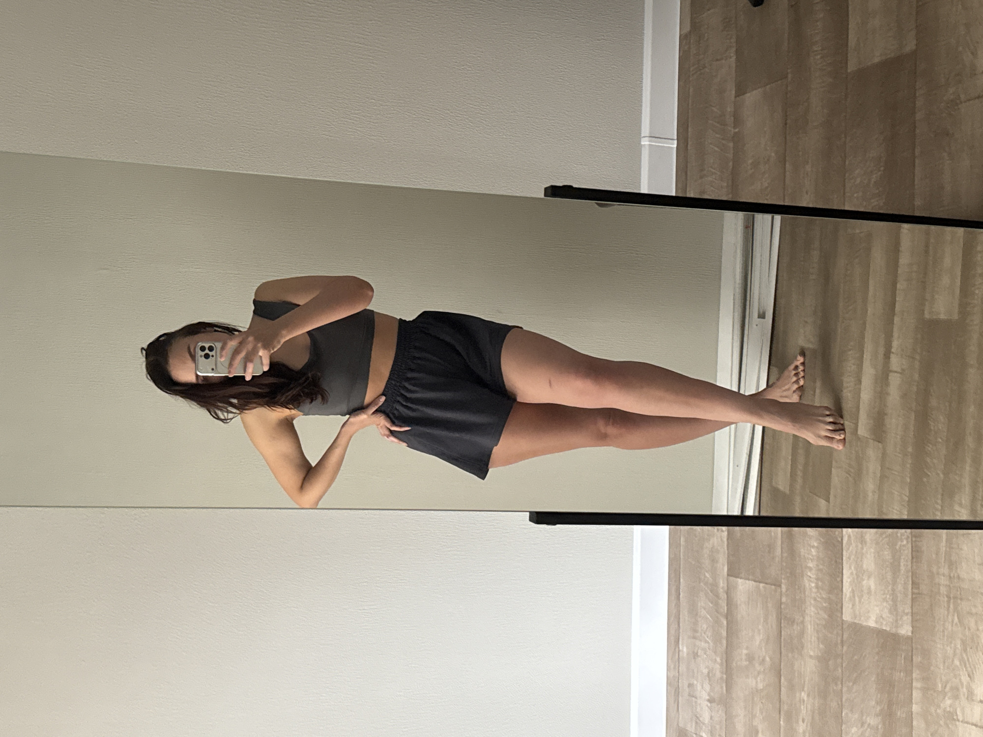 A young woman is taking a selfie in a full-length mirror. She is wearing a dark grey sports bra and matching shorts. She is standing on a wooden floor, and the background consists of a light-colored wall. The mirror is framed in black. The location is unlikely to be Chaikovsky, Russia, given the style of the interior.