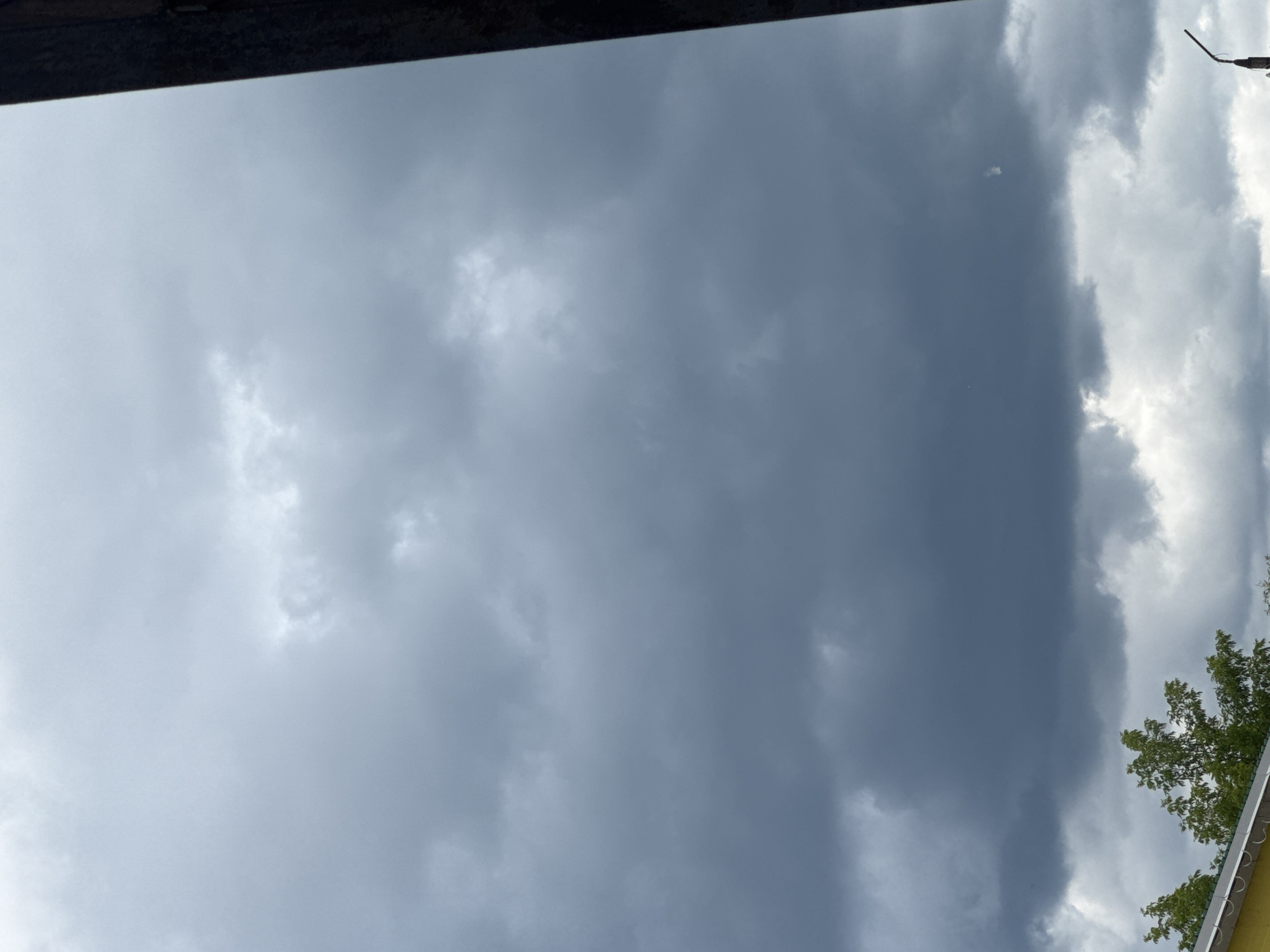 The image depicts a cloudy sky above the city of Zaokskiy, Russia. The sky is filled with dark, heavy clouds, suggesting an impending storm or overcast weather. In the lower portion of the image, the tops of green trees are visible, along with a section of a yellow building with a white roof edge. A dark vertical object, possibly a utility pole or building edge, is visible on the right side of the frame. The overall scene is muted in color due to the cloudy conditions.