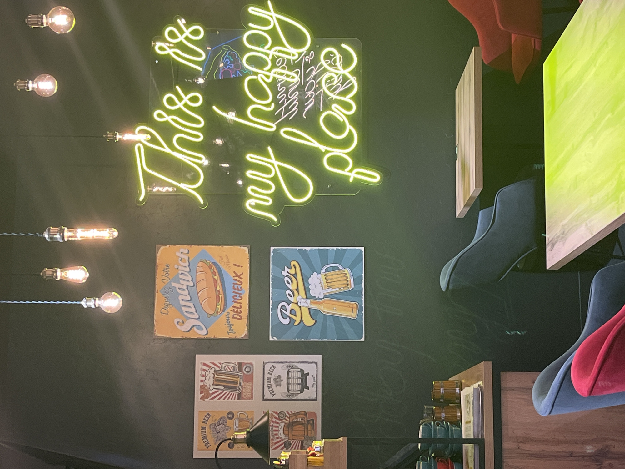 The image depicts a wall adorned with decorative elements and lighting fixtures, likely within a commercial establishment in Izhevsk, Russia. A neon sign in the upper section displays a cursive phrase in a vibrant yellow-green hue, mounted on a clear backing. Below, three rectangular signs are arranged: one featuring a sandwich illustration with French text, another showcasing a beer-themed design, and a third composed of smaller beer-related graphics. A desk lamp illuminates the lower portion of the wall. Edison-style light bulbs suspended from the ceiling add to the ambient lighting. The photograph’s composition includes a partial view of tables and chairs, indicating an interior space. The overall impression suggests a bar or restaurant setting.