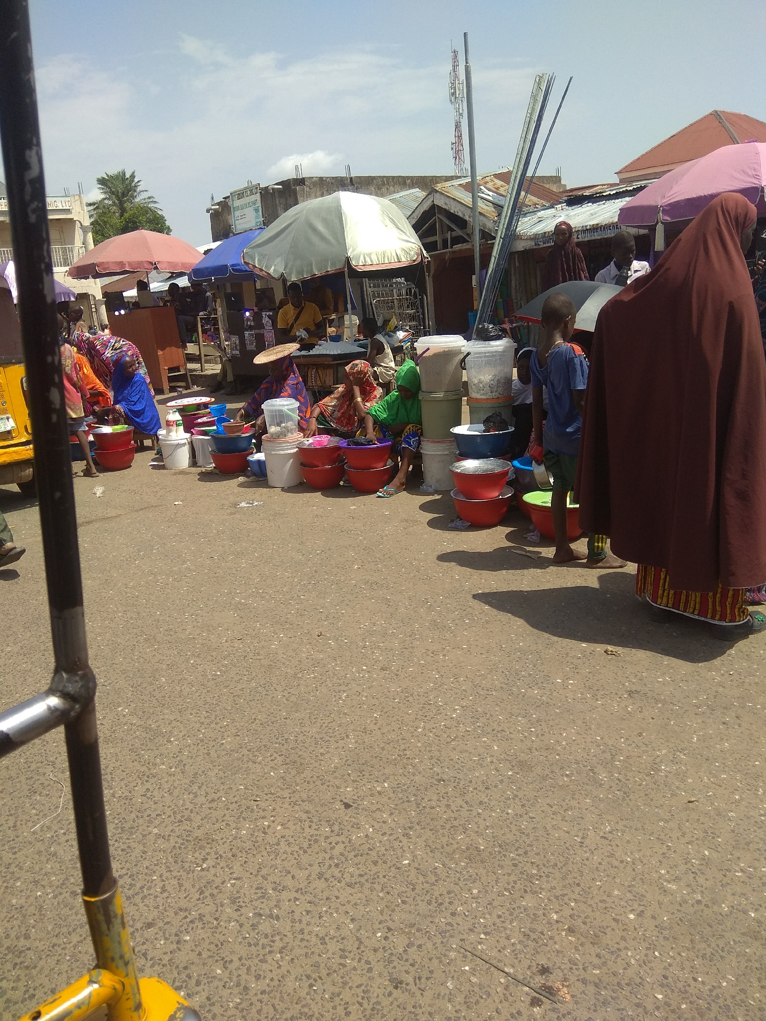 The image depicts a bustling outdoor market scene in Jalingo, Nigeria. In the foreground, a yellow and metal structure, likely part of a vehicle or cart, partially obscures the view. The ground is paved asphalt, with scattered debris. A variety of individuals, including women in colorful attire and children, are engaged in commerce. Numerous plastic bowls of various sizes and colors are displayed, suggesting a sale of household items or produce. Some individuals are interacting with the goods, while others are observing. In the background, market stalls with awnings and umbrellas provide shade, and buildings indicative of a commercial area are visible. The sky is partly cloudy, suggesting daytime. A cell tower is also visible in the distance.