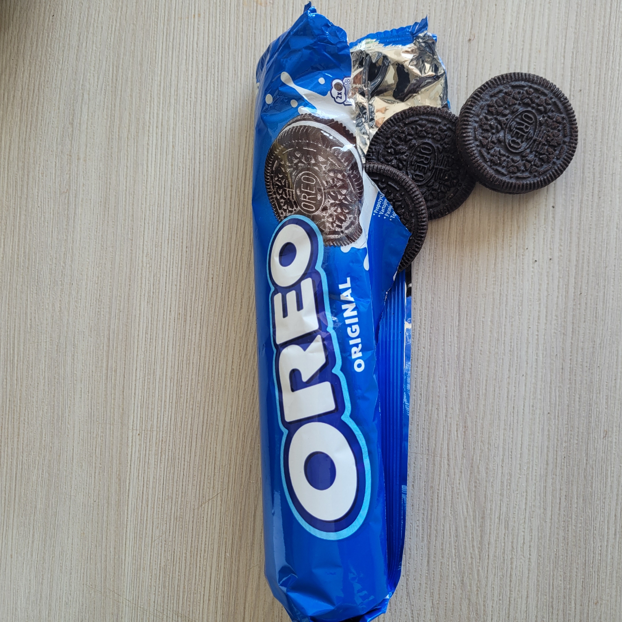 The image displays a package of Oreo Original cookies lying on a light-colored wooden surface. The blue packaging is prominent, featuring the Oreo logo and the word "Original." Two cookies are visible outside the package, one partially detached from the wrapper and another fully separate. The cookies are dark brown with a cream filling, showing the characteristic Oreo pattern. The background is a simple wooden texture. There is no indication of the city of Jūrmala, Latvia, in the image.