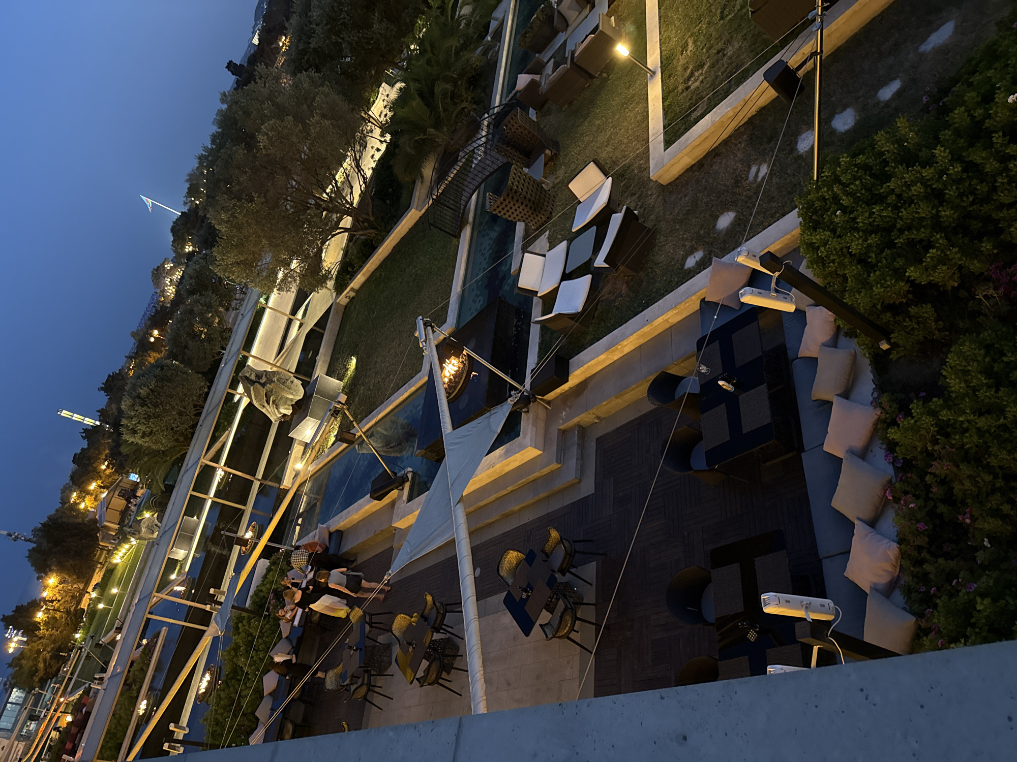 The image depicts an outdoor seating area of a restaurant or cafe in Baku, Azerbaijan, at dusk. The scene is elevated, offering a view of tables and chairs arranged on a tiled patio and a grassy area. Several groups of people are seated at tables, some engaged in conversation. A covered seating area with cushions is visible in the foreground. In the background, there are trees, outdoor lighting, and some distant structures, including what appears to be a tower. The overall ambiance suggests a social gathering at a leisure establishment.