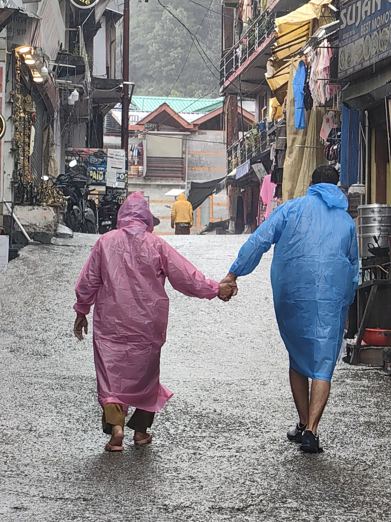In Vashisht, India, two individuals, seen from behind, walk hand-in-hand down a wet, cobbled street during a rain shower. The person on the left wears a bright pink hooded raincoat and brown pants, with one foot bare and the other in a pink sandal. The person on the right is clad in a blue hooded raincoat and dark athletic shoes. The street is lined with multi-story buildings displaying shops and goods, including awnings, clothing racks, and signage. In the background, a person in a yellow jacket walks away, and more buildings and trees are visible. The wet cobblestone street reflects the ambient light, indicating continuous rainfall. The scene captures a moment of shared experience amidst the inclement weather in this bustling Indian town.