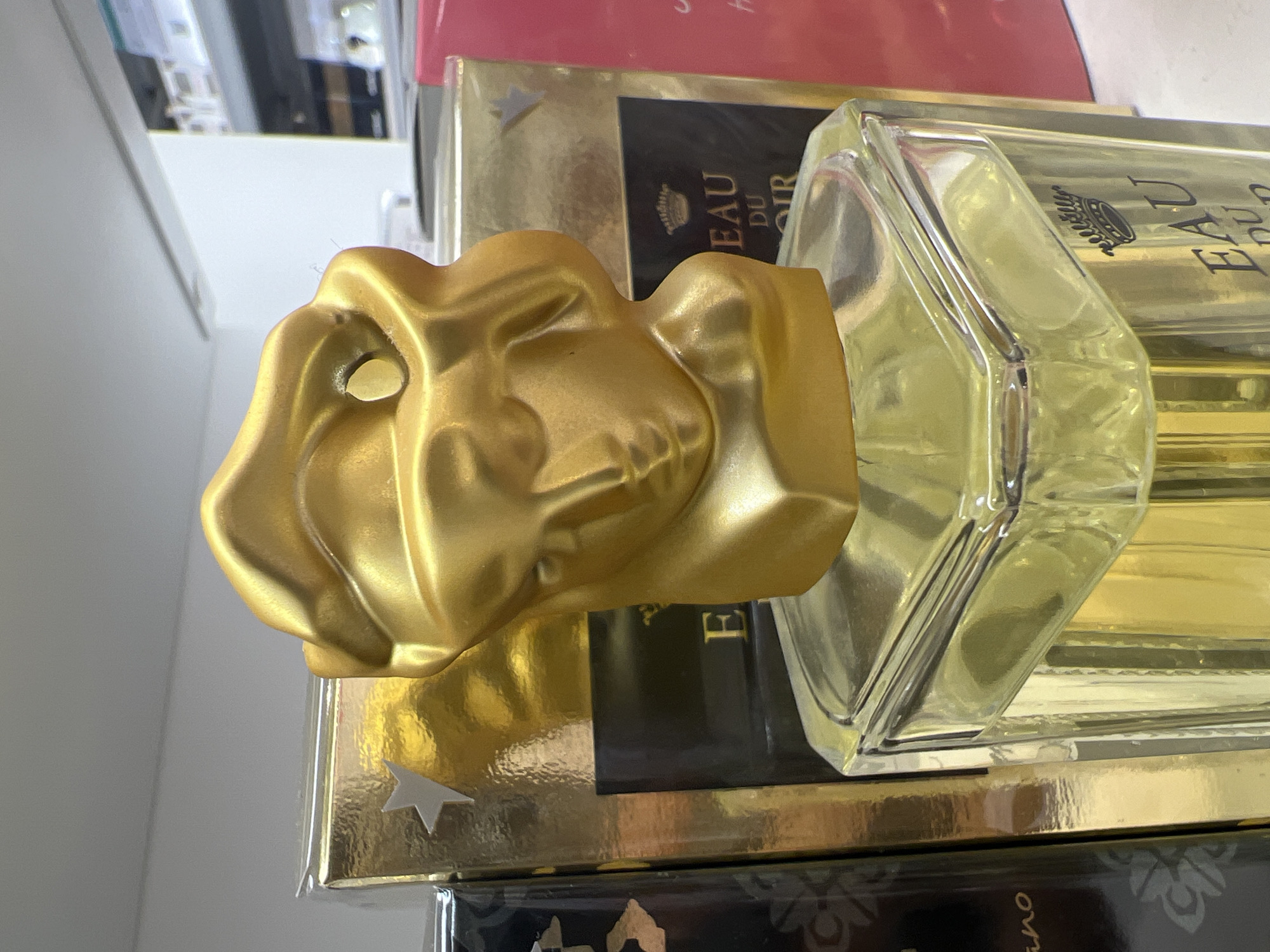 A close-up, eye-level shot showcases a distinctive perfume bottle with a striking gold sculptural lid resembling a face. The bottle is clear glass, filled with a pale yellow liquid, and has a faceted octagonal shape. Text in black on the bottle reads "EAU DU NOIR" with a small crown emblem above it. The bottle sits in front of a gold-colored box that also features a crown and text. To the left of this box is a dark rectangular box with a faint floral pattern and partial text. To the right, a bright pink box is visible. The background consists of a white shelf or wall and what appears to be a retail environment with shelves and products further back. The scene is indoors, well-lit, and the focus is sharp on the perfume bottle and its immediate surroundings. The image does not contain any information that would indicate its location within the city of Sofia, Bulgaria.