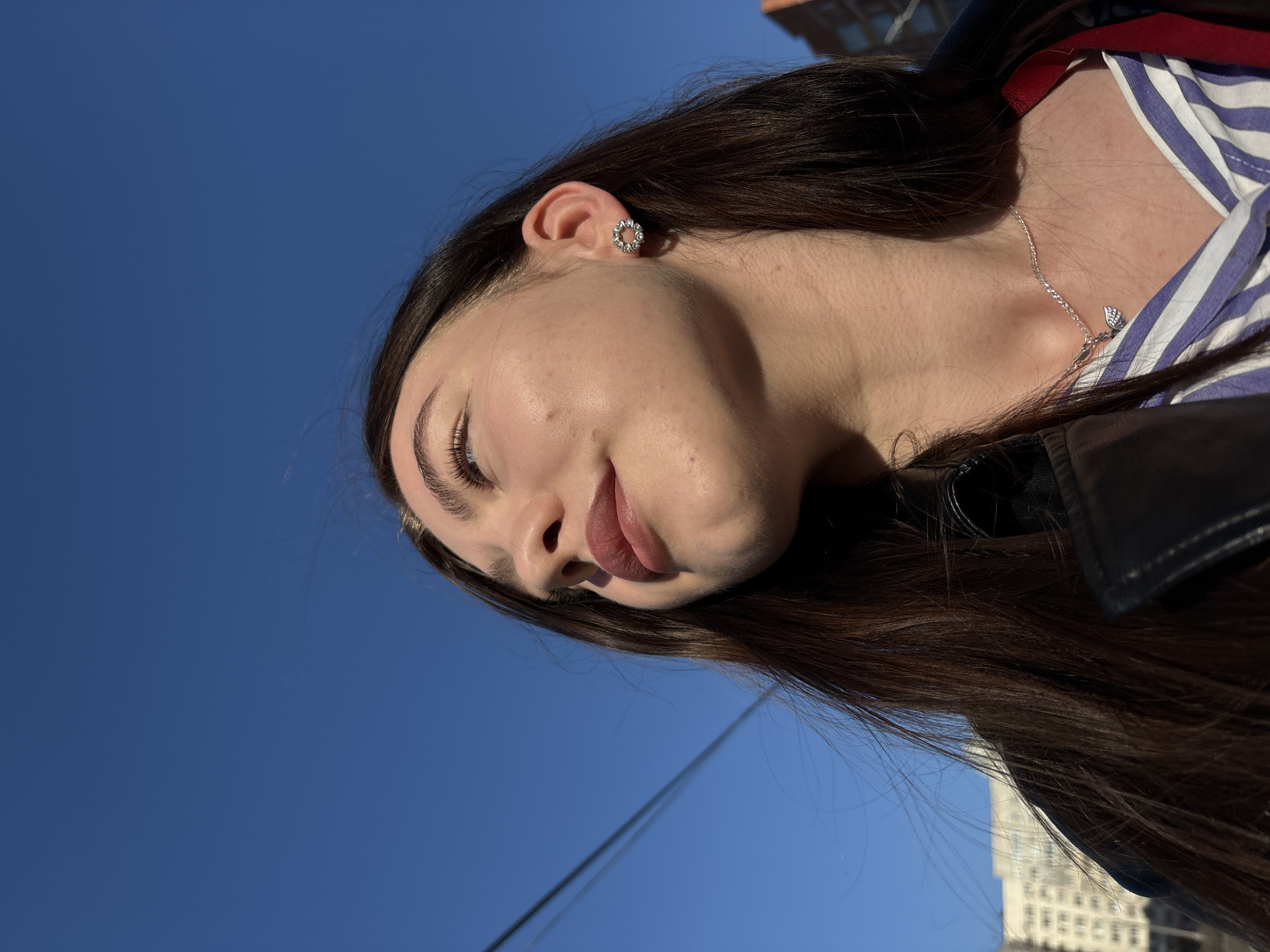 An upward-facing, close-up portrait captures a young woman from the chest up, set against a clear, deep blue sky. The woman has long, dark brown hair, parted to the side, and fair skin with visible pores and faint freckles. Her eyes are closed or looking downward, and her lips are full with a natural, rosy tone, suggesting a peaceful or contemplative expression as she basks in strong sunlight.
She wears a light-colored striped top, possibly blue and white, under a dark, possibly leather, jacket. A delicate silver chain necklace with a small, stylized pendant, resembling a feather or wing, rests on her collarbone. A circular, silver-toned stud earring, appearing to be set with small stones or details in a floral pattern, adorns her right ear.
The lighting creates distinct shadows across her face and neck, indicating a bright, sunny daytime, likely afternoon. The background is dominated by the unobstructed blue sky. In the lower right of the frame, a blurred, light-colored, multi-story building is faintly visible, suggesting an urban setting. A thin, dark utility wire traverses the sky in the lower left. There is no visible text. The scene is likely set in Perm, Russia.