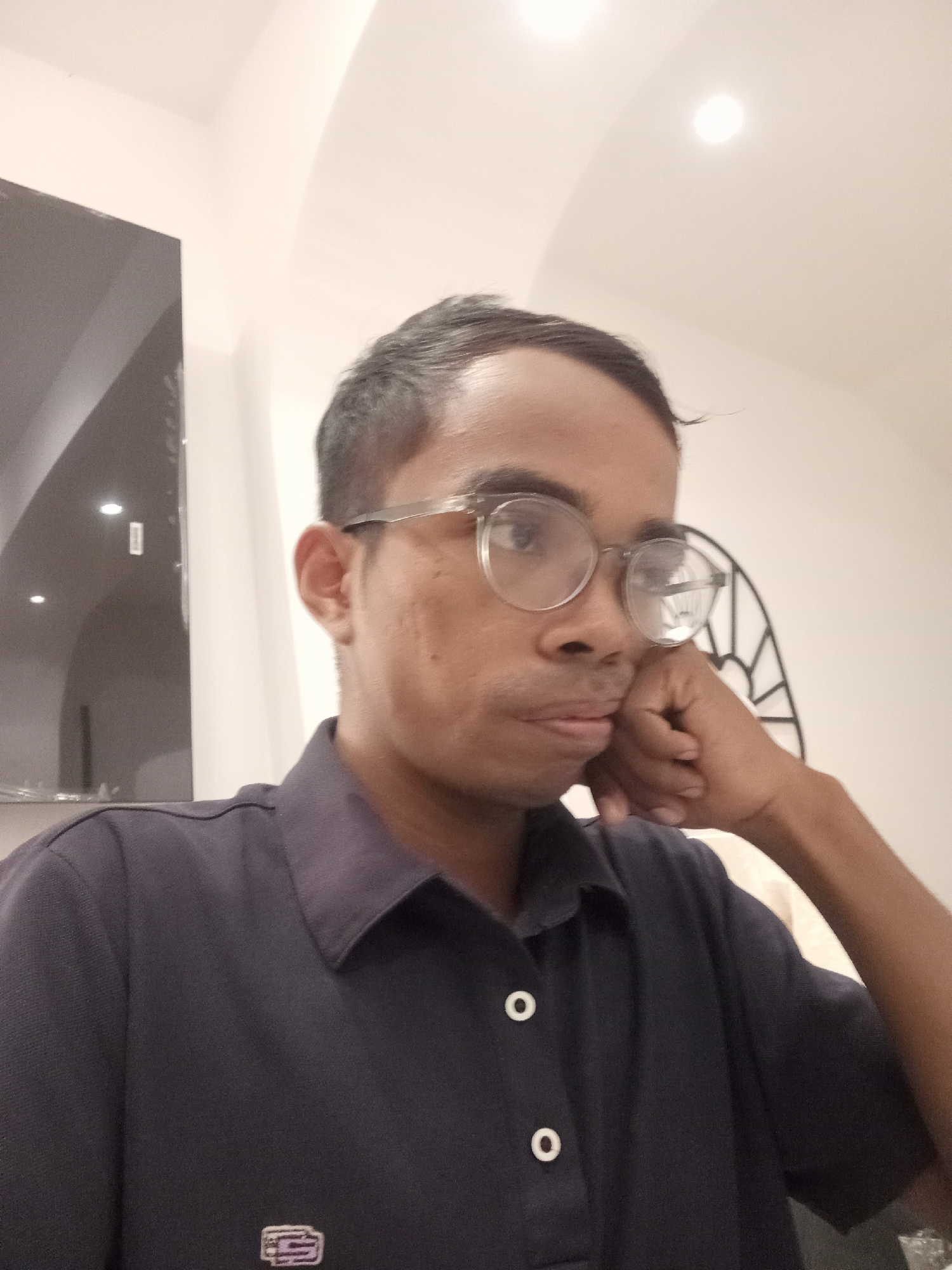 The image is a close-up, waist-up selfie of a person with dark hair, wearing glasses and a dark collared shirt with white buttons. The person's right hand is resting on their chin, and they appear to be looking away from the camera. The background is an indoor setting with a white ceiling and a dark, curved wall or structure on the left side of the frame. There is also a decorative metal object visible behind the person's head. The lighting suggests an interior environment, possibly a home or an office. The location is identified as the city of Larangan, Indonesia.