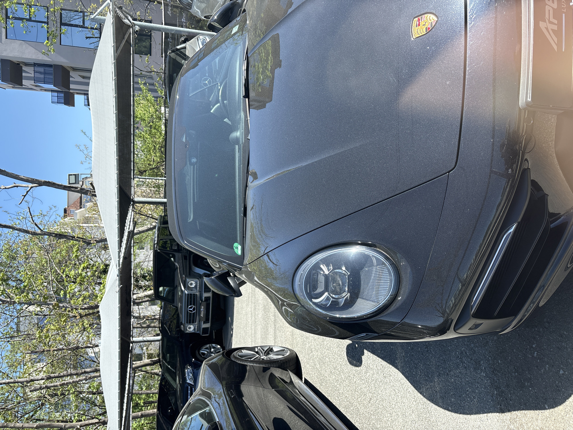 The image displays a dark gray metallic Porsche car parked outdoors on a bright, sunny day, likely at a car dealership or parking facility in Sofia, Bulgaria. The Porsche is the primary subject, captured from its front-right angle, showcasing its prominent round headlight, front bumper, and dusty hood.
The iconic Porsche crest emblem is visible on the front hood. A license plate holder on the front bumper features the text "Apex One LUX". Inside the car, parts of the steering wheel are visible through the windshield, which also has a small green sticker affixed to its lower corner. The car's surface appears dusty, reflecting the sunlight.
Behind the Porsche, a black Mercedes-Benz G-Class SUV is parked, with its distinctive grille and front lights clearly visible. The Mercedes-Benz emblem and the text "Apex One" are visible on its grille. A third dark vehicle is partially visible in the foreground, to the right of the G-Class.
The cars are parked under a light-colored fabric canopy structure, which provides shade. Beyond the canopy, trees with green leaves and a clear blue sky are prominent, suggesting pleasant, clear weather. In the background, modern buildings with several reflective windows can be seen. The ground is asphalt, and sharp shadows are cast by the vehicles, further indicating strong daylight. No people are visible in the scene.
