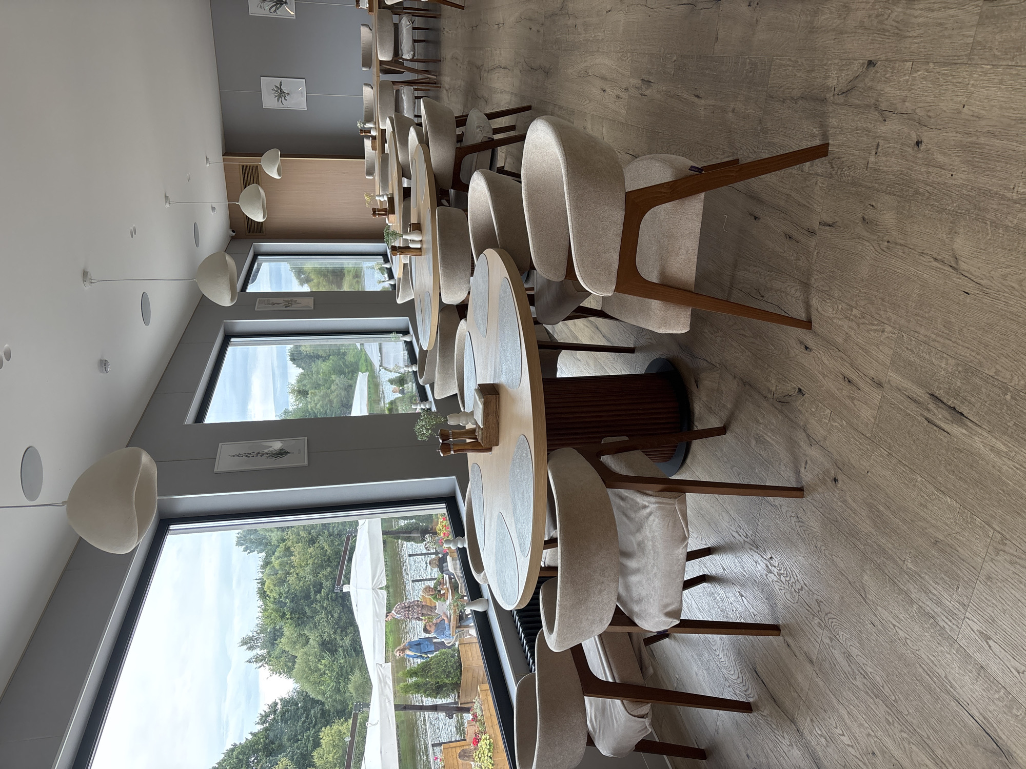 The image depicts the interior of a restaurant with large windows overlooking a body of water and trees, likely located in Zvenigorod, Russia. The restaurant features multiple round dining tables with light-colored chairs. The tables are set with placemats, salt and pepper grinders, and small decorative vases with greenery. The chairs are upholstered in a light beige fabric and have wooden frames. In the background, through the windows, several white umbrellas are visible, suggesting an outdoor seating area. Some people are visible outside, near the water, including two adults and a child sitting at a table. The interior walls are painted a neutral gray, and the floor is a light wood laminate. Several pendant lights with a dome shape hang from the ceiling. The overall ambiance is calm and contemporary.