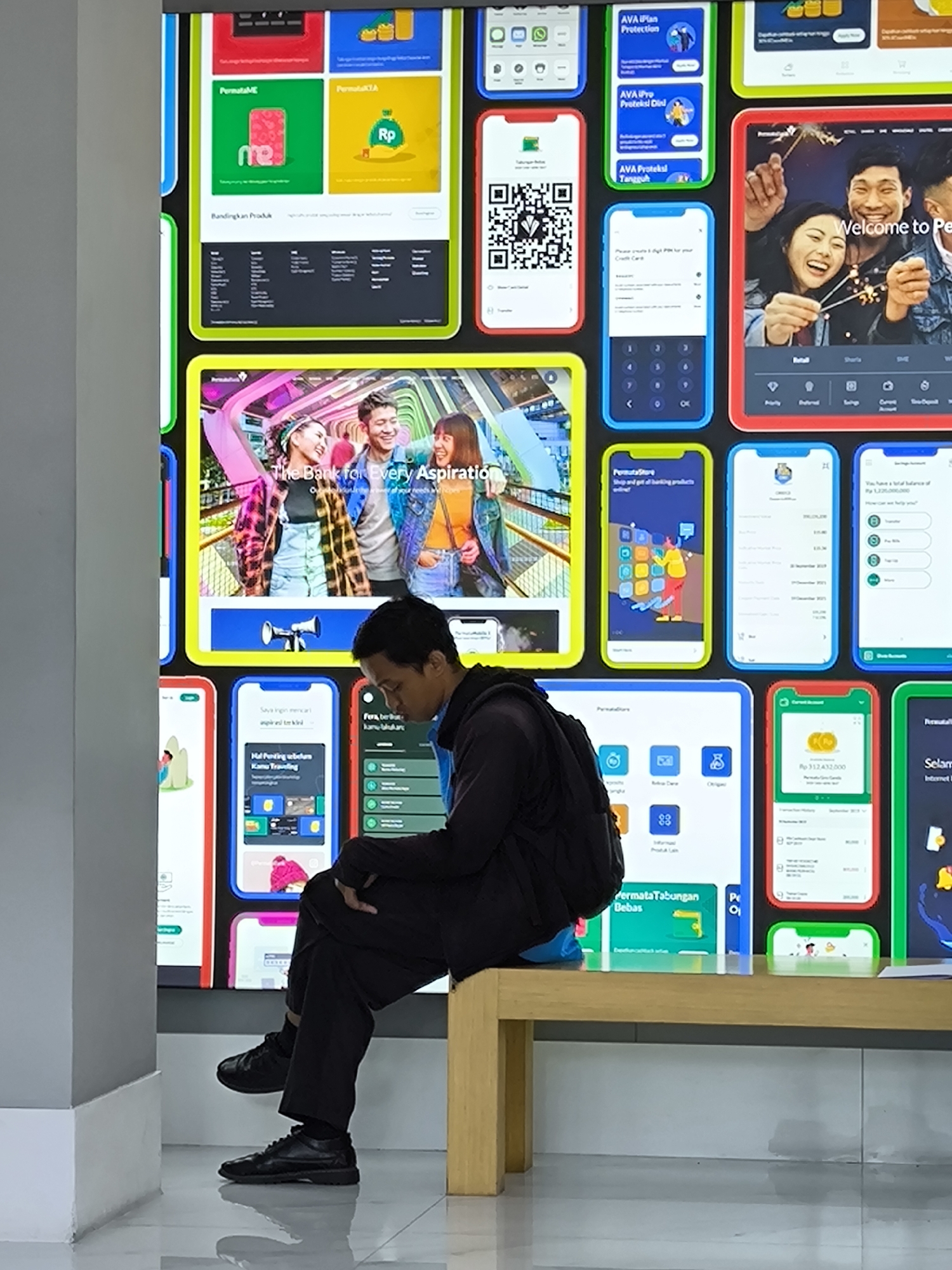 The image shows a man sitting on a light-colored wooden bench in front of a large digital display composed of smaller screens. The screens depict mobile phone app interfaces with Indonesian text and imagery. The man is dressed in dark clothing and appears to be looking down. The location seems to be an indoor public space with a modern aesthetic, featuring a gray wall to the left and a tiled floor. The environment suggests a location in North Kuta, Indonesia based on the text and brand names on the screens.