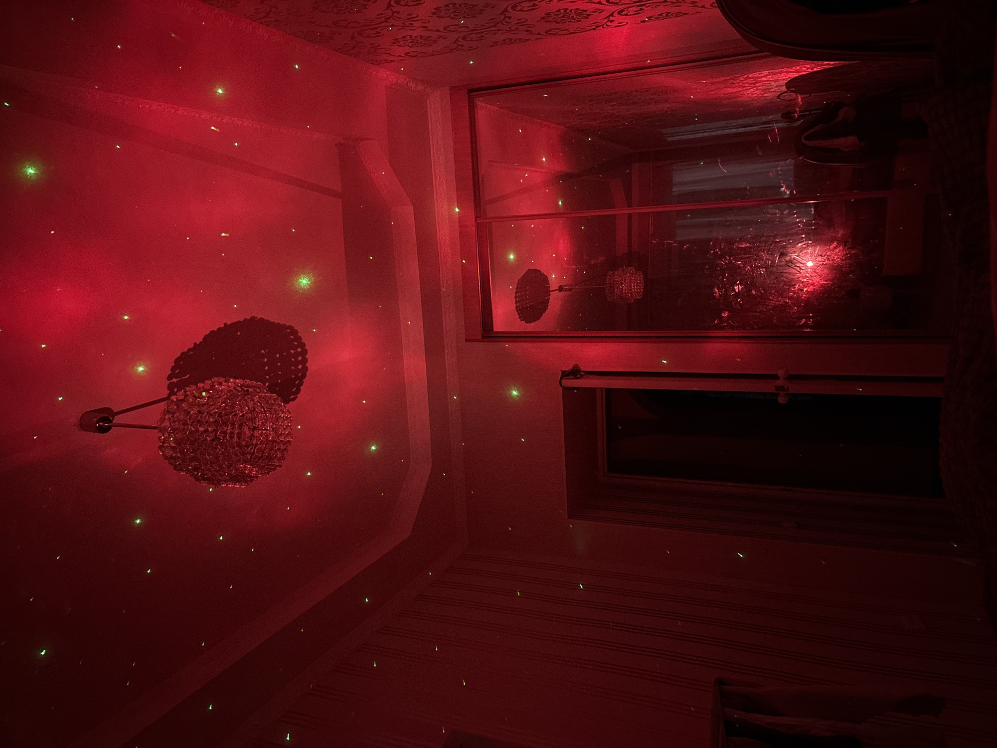The image captures an interior room in Veliky Novgorod, Russia, bathed in pervasive red ambient light and speckled with numerous small green laser points across the ceiling and walls. A chandelier, comprising two spherical clusters of reflective elements, hangs centrally from the white, crown-molded ceiling. The left wall features striped dark red wallpaper, contrasting with the floral-patterned dark red wallpaper on the right wall. A large, mirrored wardrobe occupies the right side of the room, extensively reflecting the chandelier, green laser points, and the red illumination. A dark, open doorway is visible on the left side of the room. The immediate foreground reveals a dark, textured surface, possibly carpeting, with a partial view of a dark, curved object on the extreme right.