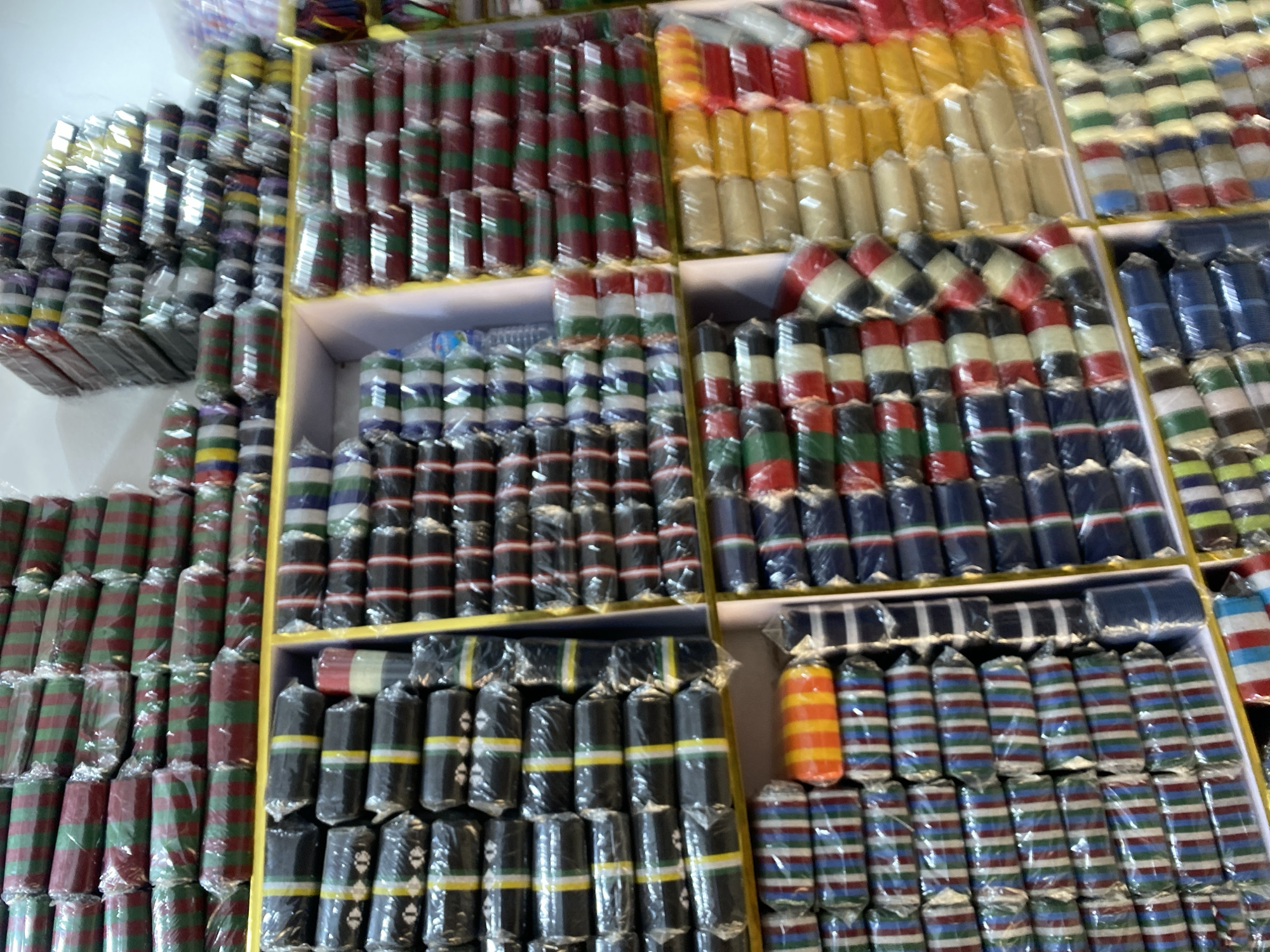 The image shows shelves in a shop, potentially in Bukuru, Nigeria, stacked with rolls of what appear to be colorful fabrics or ribbon, possibly packaged in clear plastic. The rolls are arranged in rows and columns on the shelves, with a variety of color combinations, including red and green, yellow, blue, black and white, and multicolored stripes. The overall appearance suggests a retail environment specializing in fabrics or crafting supplies.