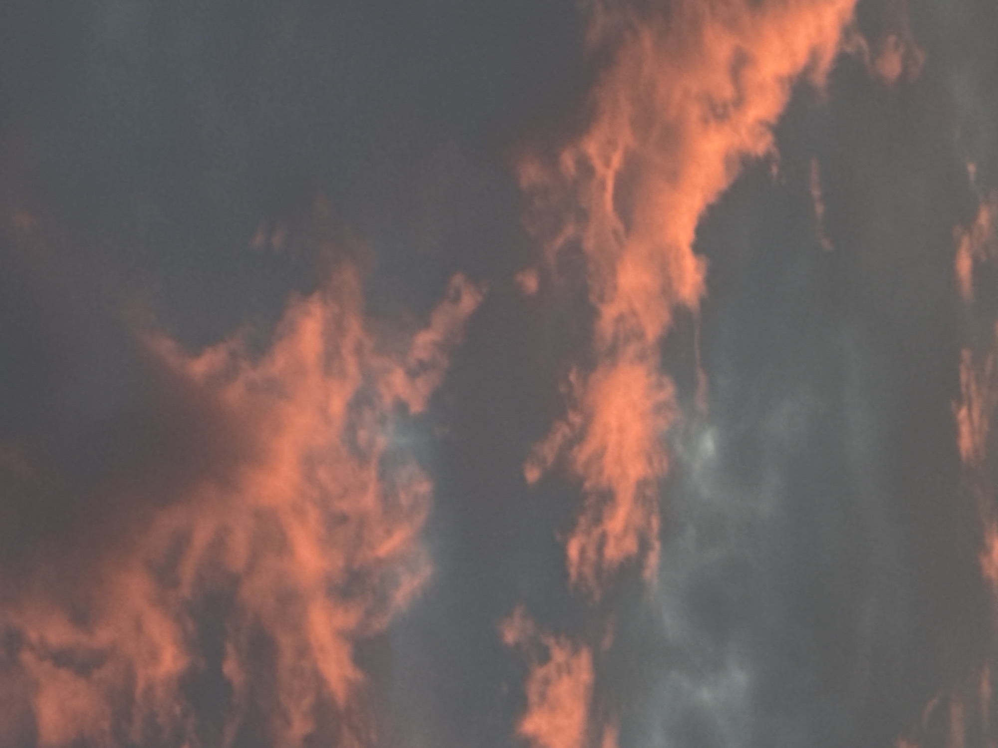 The image is a close-up, vertically oriented, low-angle shot of a sunset sky filled with clouds. The clouds are illuminated with a vibrant orange and pink hue, contrasting with the darker, greyish-blue of the surrounding sky. The scene is an outdoor environment. There are no visible people, subjects, objects, actions, or interactions. The image captures a natural phenomenon.