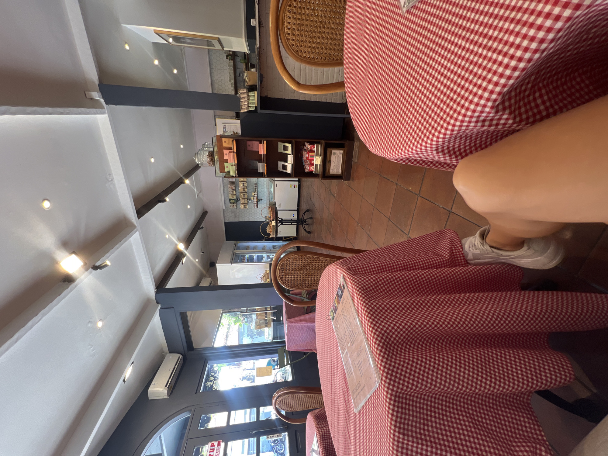The image shows the interior of a café in North Kuta, Indonesia. The café has tables covered with red and white checkered tablecloths. The tables are accompanied by rattan chairs. A person's legs and feet wearing white sneakers are visible at a table. A menu stand is placed on one of the tables. The café's interior features dark grey structural supports, white walls, and a high ceiling. A shelving unit with displayed items, refrigerators, and other furnishings are present. The entrance has glass doors and windows.
