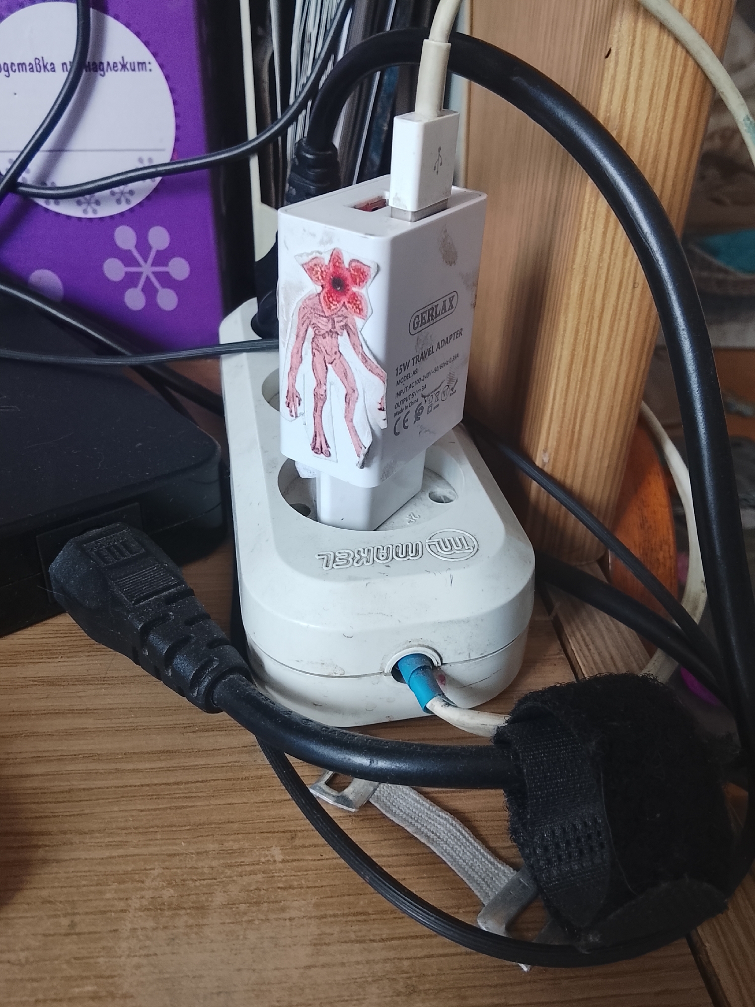 The image depicts a close-up view of a power strip with a wall adapter plugged into one of its sockets. The wall adapter is white and has a sticker of a creature from "Stranger Things" attached to it. The adapter is labeled "GERLAX 15W TRAVEL ADAPTER". The power strip itself is also white and appears to be a multi-socket unit. A black power cord with a thick connector is plugged into the power strip and extends out of the frame. Several other black cables are also visible, some coiled and secured with a black velcro strap. The scene is set on a wooden surface. A portion of a purple object with white snowflake-like designs is visible in the upper left corner of the image, with some text in Cyrillic script that reads "поставка на надлежим:". The background shows a wooden structure and out-of-focus objects. There are no people visible in the image. The overall scene suggests a domestic or office environment where electrical devices are being powered. The presence of the Cyrillic text might suggest a location in a country that uses this script, such as Kazakhstan, where Almaty is a major city.
