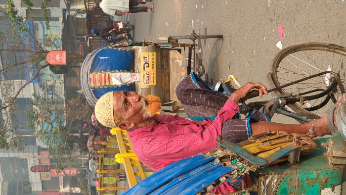 An elderly man, identified as a rickshaw puller, is seated on his blue and green bicycle rickshaw in a street in Dhaka, Bangladesh. He wears a cream-colored kufi and a pink long-sleeved shirt, with a dark striped lungi. His beard and mustache are dyed a distinctive orange-red hue. His hands rest on the rickshaw's handlebars, and he wears flip-flops.
In the mid-ground, a second bicycle rickshaw with a blue canopy is visible, bearing a "DOT INTERNET" advertisement in Bengali and English. To the left, yellow metal barricades separate the street from a sidewalk area where several individuals are present. To the right, a pedestrian in a light-colored shirt walks away from the camera, and a motorcyclist in a blue helmet is visible further back. The background features multi-story urban buildings, one displaying a red sign with a tree logo (Akij). Sparse trees are interspersed along the street. The asphalt roadway contains minor debris. The scene is illuminated by daylight, with a visible atmospheric haze typical of a busy urban environment. No specific interactions or significant actions are observed beyond passive presence and movement.