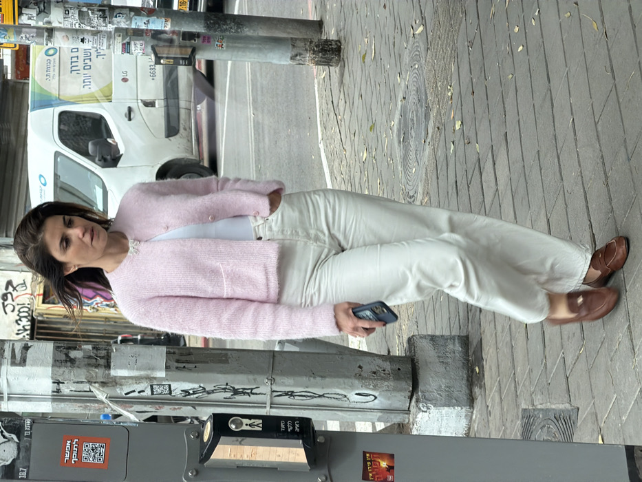 An adult female is captured mid-stride, walking from left to right on a paved city sidewalk in Tel-Aviv, Israel. She has dark brown, shoulder-length hair, styled with a slight wave, and a few strands appear to be blowing in the wind. She wears a light pink, fuzzy, cropped cardigan with two small front pockets and a white undershirt. A silver-colored bow-shaped brooch is pinned to her left lapel. Her lower attire consists of light-colored, high-waisted, wide-leg pants and brown leather loafers with a gold-colored metallic buckle. Her left hand holds a dark blue smartphone, while her right hand is in her pants pocket. Her expression is neutral, with her gaze directed slightly downward.
To her left, two grey metal poles stand on the sidewalk. The pole closest to the viewer features a black pedestrian crossing signal box with a yellow/orange indicator light, and a white sign with a QR code and Hebrew text. Below it, a red and black sticker with Hebrew text is visible. The second pole is thinner and covered in multiple stickers and some faded text. Behind the poles, a wall shows dark graffiti. The sidewalk is paved with rectangular light grey/beige pavers, with scattered dry leaves and debris. A circular metal manhole cover is visible on the right side of the sidewalk.
In the background, beyond the sidewalk and an asphalt road, a white commercial van is parked or moving, displaying light blue and green graphics, Hebrew text, an "LG" logo, and a phone number (+6683...). Part of a dark-colored vehicle is visible behind the van. The overall scene depicts a typical urban street environment under daylight conditions.