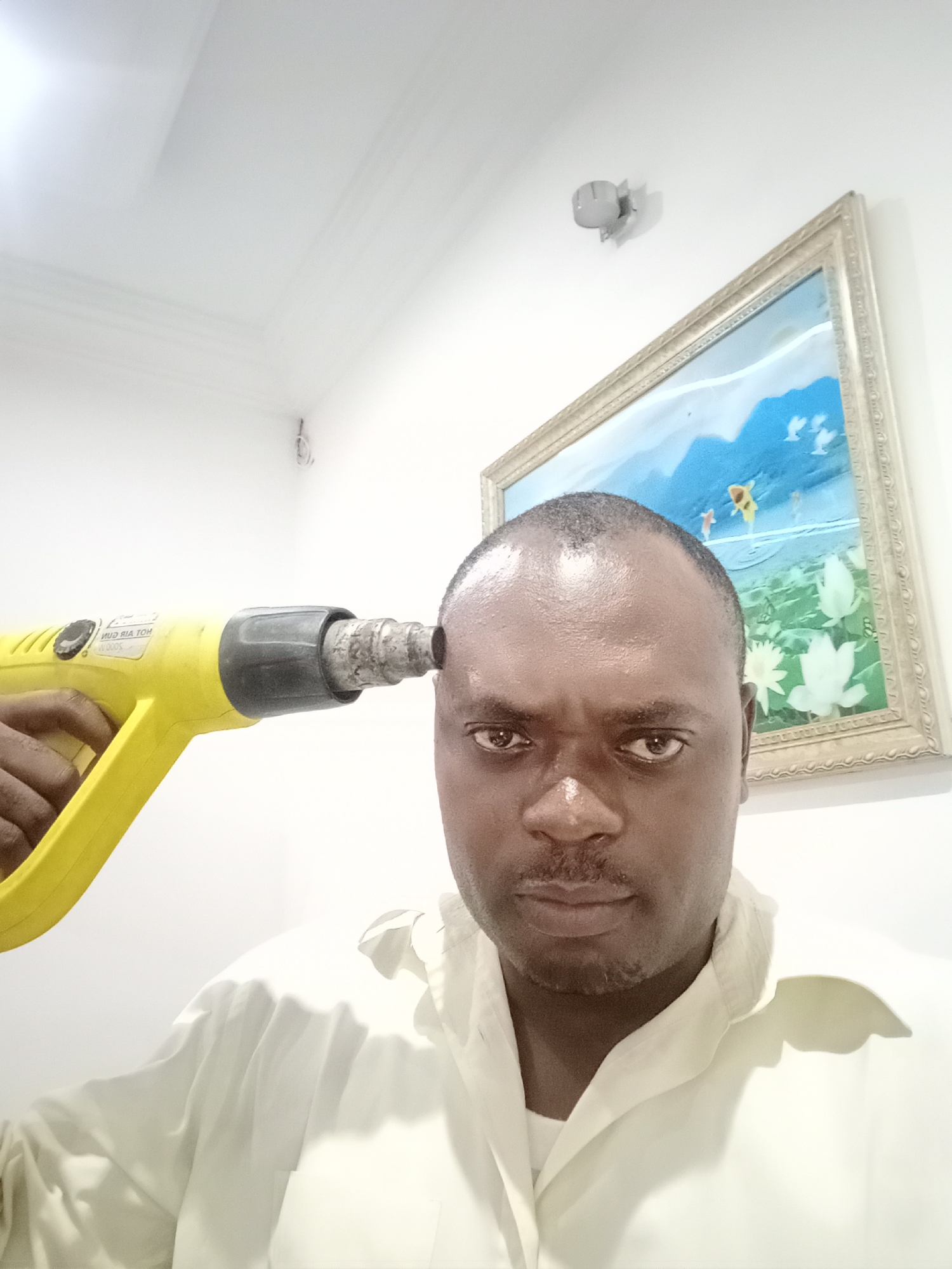 A man is holding a yellow heat gun to his forehead. He is wearing a light-colored shirt. Behind him is a painting in a gold frame and a light fixture on a white wall. The location is Gwarinpa, Nigeria.