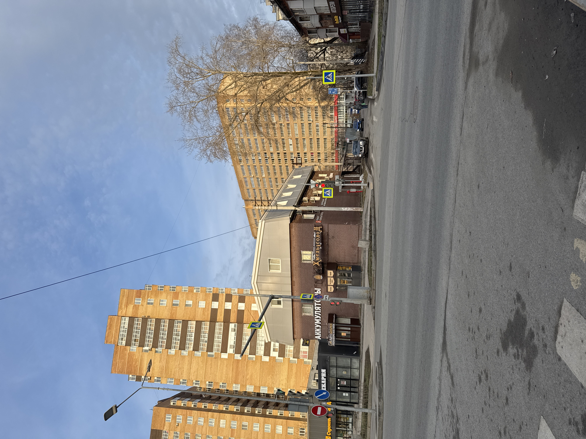 The image shows a street scene in Perm, Russia. The perspective is from street level, looking down the asphalt road. The sky is blue with scattered clouds.
To the left are two tall apartment buildings constructed from light brown brick, with rows of windows. Power lines stretch across the top of these.
On the right, buildings line the street. A smaller, multi-level brick building is visible, bearing signage, including "АККУМУЛЯТОРЫ", indicating an automotive business, and a sign for a beer establishment. The storefronts are in close proximity to a pedestrian crosswalk with visible yellow road signs. Farther up the road is a larger building in a similar brown color. There is a mature, leafless tree in front of it. Cars, including a dark-colored sedan, are parked along the curb.