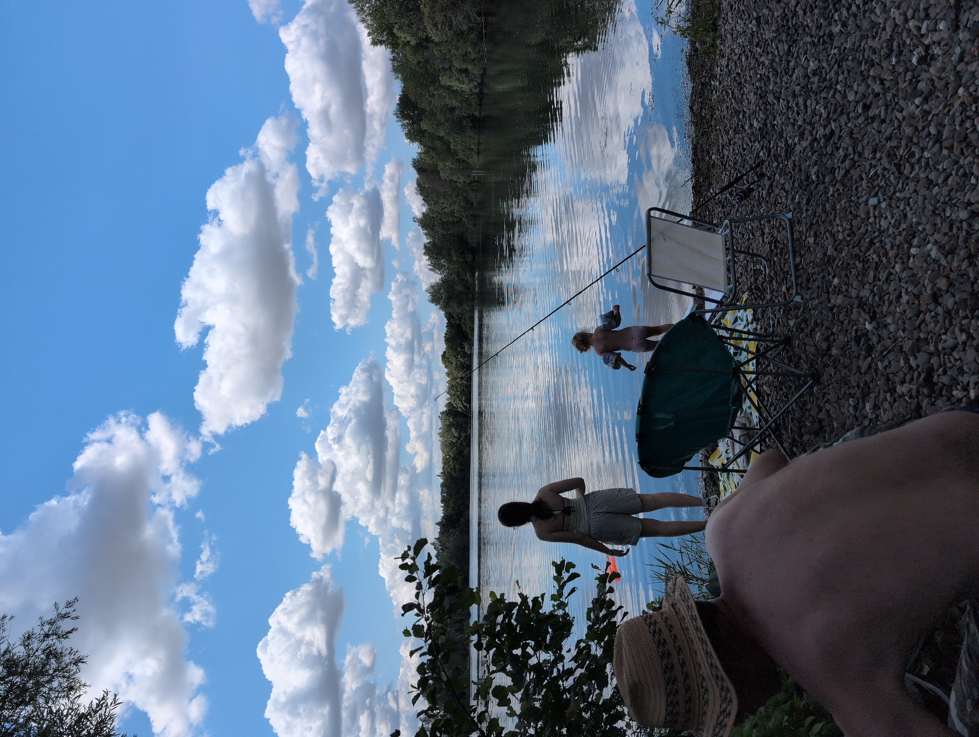 The image depicts a serene lake scene in Petershagen, Germany, on a bright, cloudy day. The sky is a vibrant blue, adorned with fluffy white clouds that cast soft shadows on the water. The lake itself is calm, with reflections of the sky and surrounding trees rippling on its surface.
In the foreground, a man wearing a straw hat is seen from behind, his back bare, enjoying the view. To his right, two camping chairs are set up on the pebbly shore, one a dark green and the other a lighter beige. A fishing rod leans against the beige chair.
Further back, closer to the water's edge, a woman stands facing the lake, wearing a striped tank top and shorts. She holds a bright orange object. A young child, wearing arm floaties, stands in the shallow water, also facing the lake. The shoreline is lined with lush green trees, creating a natural backdrop. The overall impression is one of peaceful outdoor recreation.