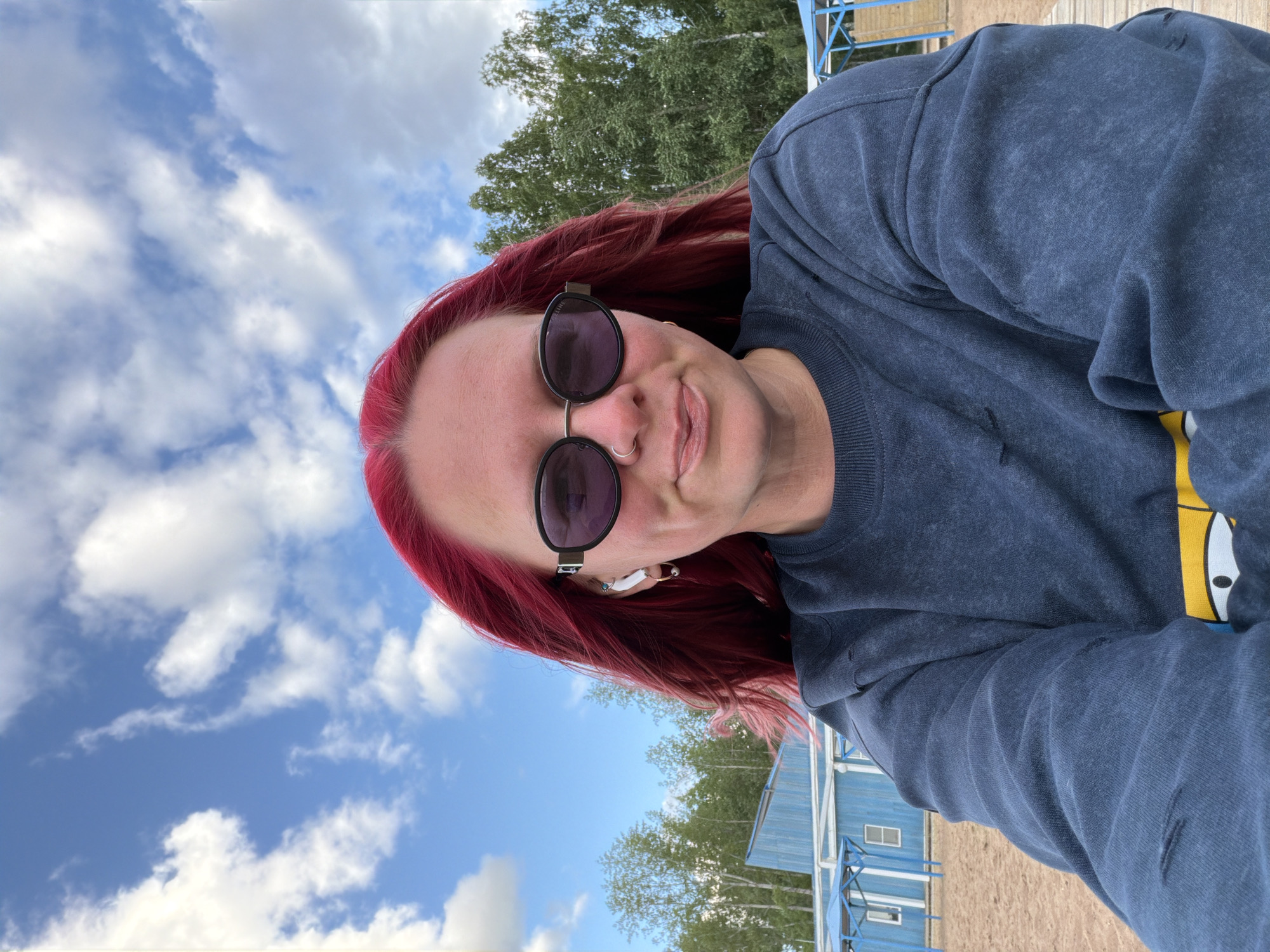 In Chaikovsky, Russia, a woman with bright pink hair and sunglasses takes a selfie outdoors. She is wearing a dark blue sweatshirt with a cartoon design visible on the lower part of the garment. She has earbuds in her ears and a nose ring. The background shows a sandy area, trees, a blue building, and a sky with scattered clouds.