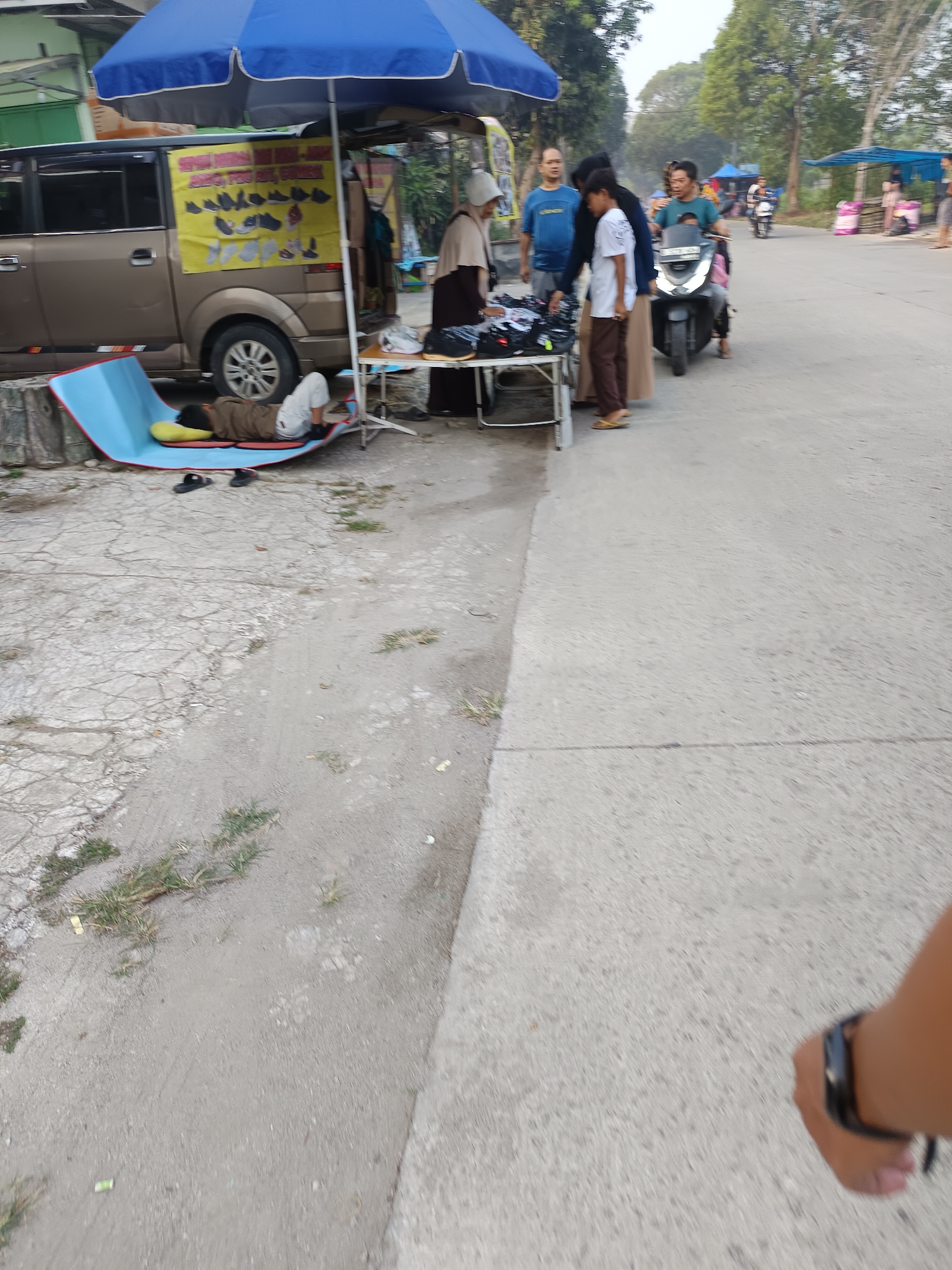 This image captures an outdoor market scene in Sodong, Indonesia, under a large blue umbrella. A person is sleeping on a blue mat near a tan van, which has a yellow banner displaying images of shoes and text in Indonesian. In front of the van, a table is set up with various clothing items displayed. Several people are gathered around the table, seemingly interacting with the goods. A man is riding a motorbike with passengers behind him, and other market stalls are visible in the background, with trees and a road forming the environment.