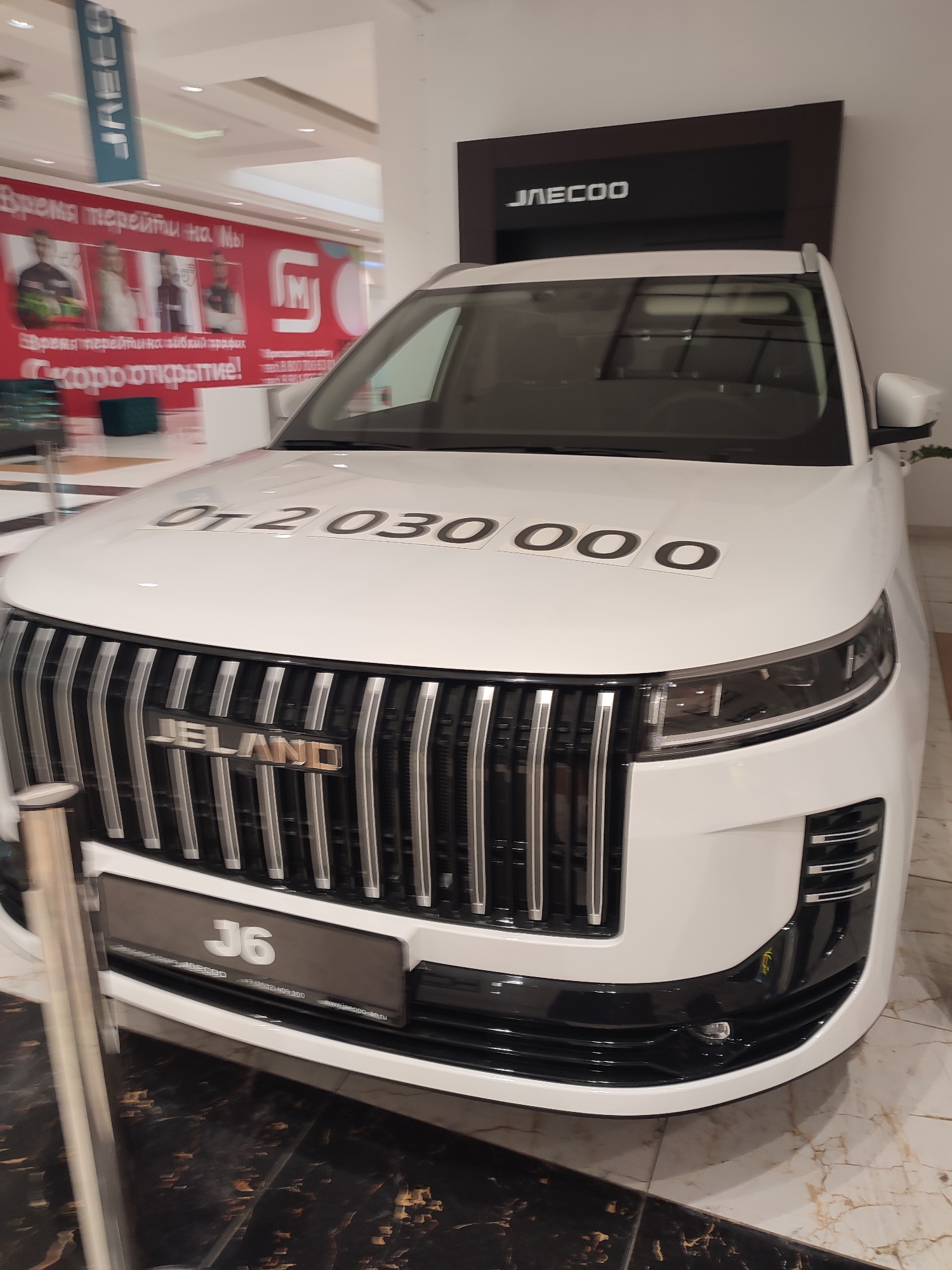 The image shows the front of a white car parked indoors, likely in a showroom or dealership. A banner with the JAECOO logo is visible above the car. On the hood of the car, the text "ОТ 2 030 000 0" is displayed on a sticker, indicating a starting price. The car's grille features the JELAND logo and the model designation "J6". A sign below the grille displays additional text, including a phone number and a website address that suggests the location is in Orenburg, Russia. The environment appears to be a well-lit indoor space with tiled flooring. No people are clearly visible in the image.
