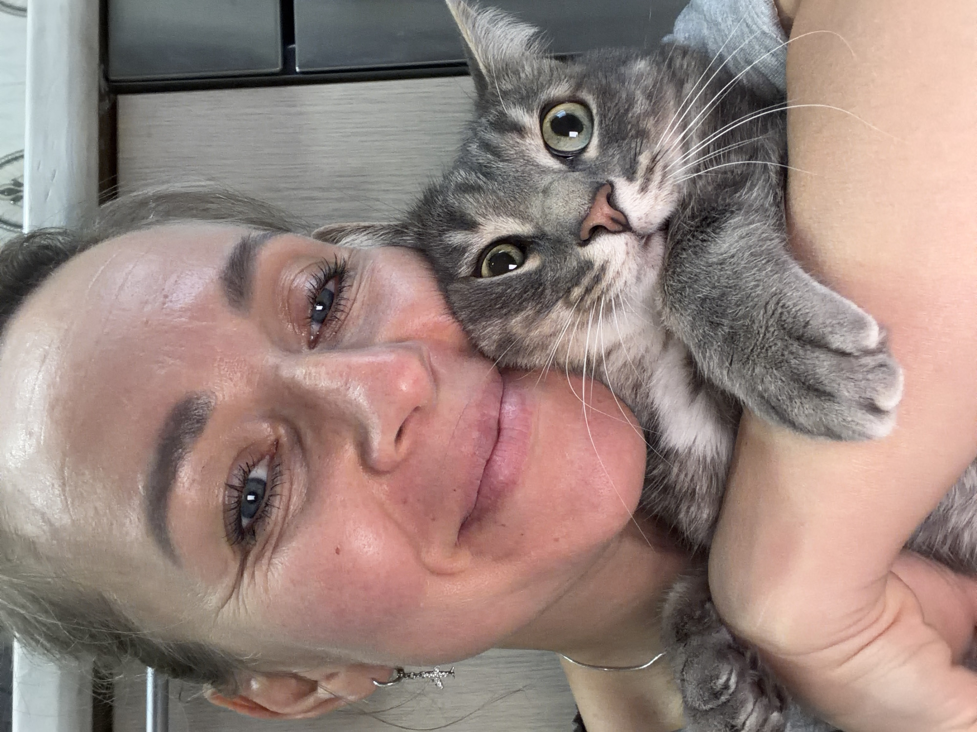 A woman holds a gray tabby cat close to her face. The woman has fair skin, blue eyes, and light brown hair pulled back from her face. She has a small earring in her left ear and wears a thin silver necklace. Her facial expression is a slight smile, with her cheek pressed against the cat. The cat has green eyes and white whiskers. The background is a combination of light gray and beige surfaces, with a section of dark gray or black visible in the upper left corner. The location is not discernible from the image.