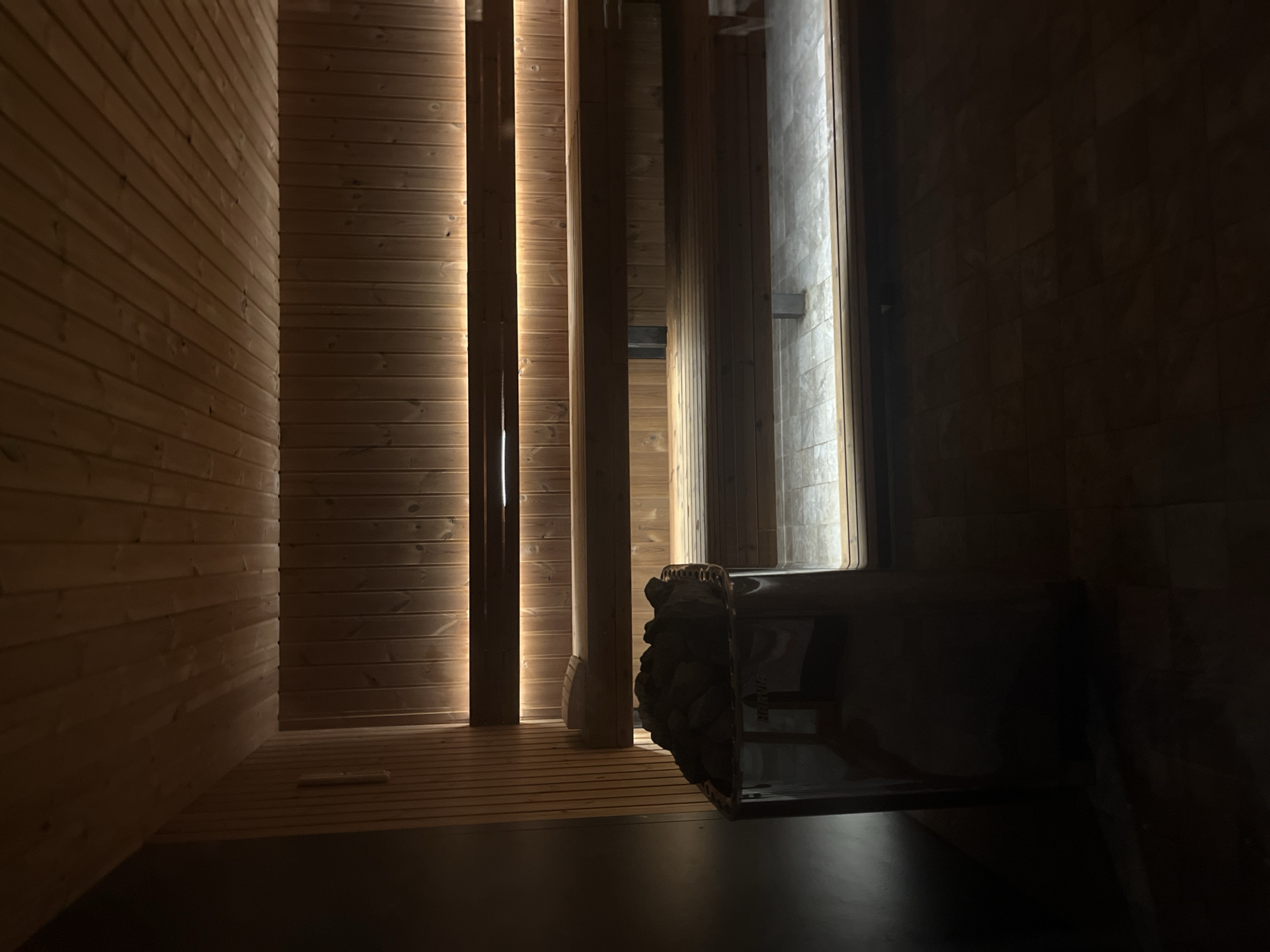 The image captures the interior of a modern sauna or steam room, characterized by its warm, subdued lighting and natural materials. The setting is dimly lit, creating an intimate and relaxing atmosphere.
The primary subjects are the architectural elements and fixtures of the sauna. The walls are predominantly clad in light-toned, horizontally paneled wood, giving the space a clean and natural aesthetic. Integrated strip lighting runs vertically along some of the wooden slats on the left wall, emitting a soft, golden glow that highlights the wood grain and provides ambient illumination. Another vertical light source is visible on the far right, illuminating a section of wall that appears to be made of textured stone or tile.
In the mid-ground on the right, a dark, metallic sauna heater is prominently featured, topped with a mound of stones, ready for use to generate steam. Below it, a dark, flat surface forms the floor or a lower bench area. Wooden benches, also constructed from horizontal slats, are visible along the left wall, typical for seating in a sauna.
The activity taking place is implied to be a relaxation or wellness session within a sauna. The time of day is not explicitly shown but the atmospheric lighting suggests evening or an enclosed environment designed for deep relaxation. No people are visible. The overall mood is tranquil and inviting. There is no visible text in the image. The location context provided is Sofia, Bulgaria, indicating this is likely part of a spa or wellness facility in that city.