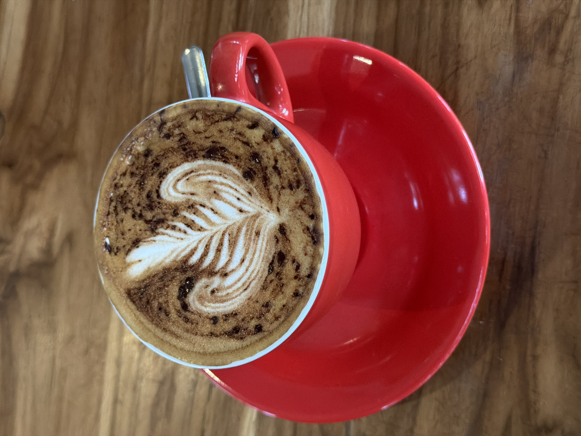 A top-down shot shows a red cup and saucer on a wooden table. The cup is filled with a cappuccino, adorned with latte art depicting a leaf-like pattern. A small silver spoon rests in the cup, next to the handle. The scene is captured in North Kuta, Indonesia.