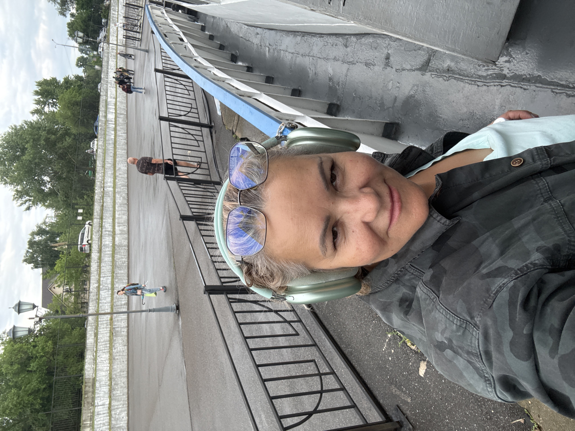 The image is a selfie taken by a woman wearing light green headphones and glasses. She is wearing a dark, camouflage-patterned jacket over a light blue shirt. The background suggests an outdoor setting with a walkway or bridge. In the distance, several people are visible walking on the path, and there are trees and lampposts. The ground appears wet, indicating recent rain. The woman has a gentle smile on her face, suggesting a relaxed or content mood. The scene is likely in Kazan, Russia, based on the provided location context. The overall impression is of a casual moment captured outdoors, possibly during a walk or while sightseeing.