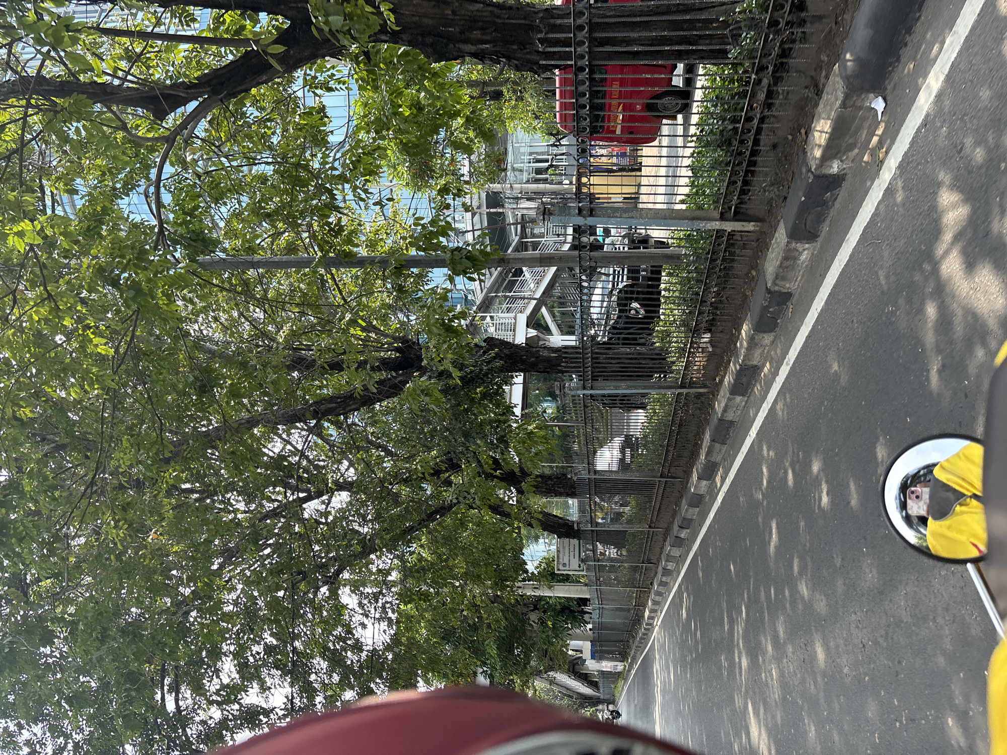 The image shows a street scene in North Jakarta, Indonesia, taken from the perspective of a vehicle, likely a motorcycle, as suggested by the side mirror in the lower right corner. The mirror reflects the photographer wearing a yellow shirt and holding a smartphone. A road stretches forward, lined with a metal fence and a row of trees on the left. Behind the fence, glimpses of buildings and an elevated pedestrian walkway are visible. A red vehicle is parked alongside. The road surface is asphalt, marked with a white line and shadows cast by the trees. The lighting suggests a sunny day.