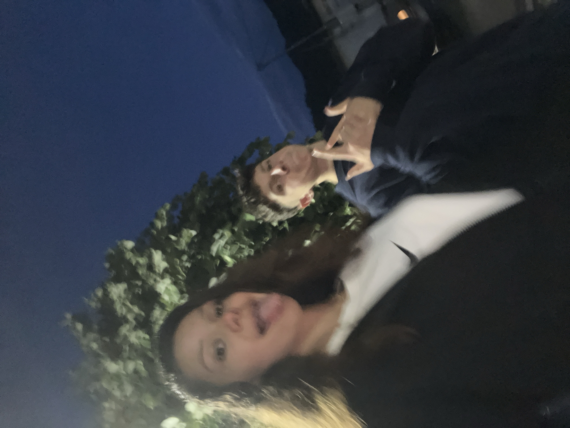 The image depicts two young individuals outdoors at night or dusk in Zlatograd, Bulgaria. On the left, a female with long dark hair and a black jacket over a white t-shirt with a visible Nike swoosh logo is captured from the chest up, looking directly at the camera with her tongue extended. To her right, a male in a dark blue hooded garment is partially obscured and out of focus, with puffed cheeks and his right hand raised, displaying a gesture with extended index and pinky fingers. The background features illuminated green foliage and a dark blue sky, with faint architectural outlines and distant topographical features visible on the right horizon. The image exhibits a shallow depth of field and some digital noise, consistent with low-light photography.