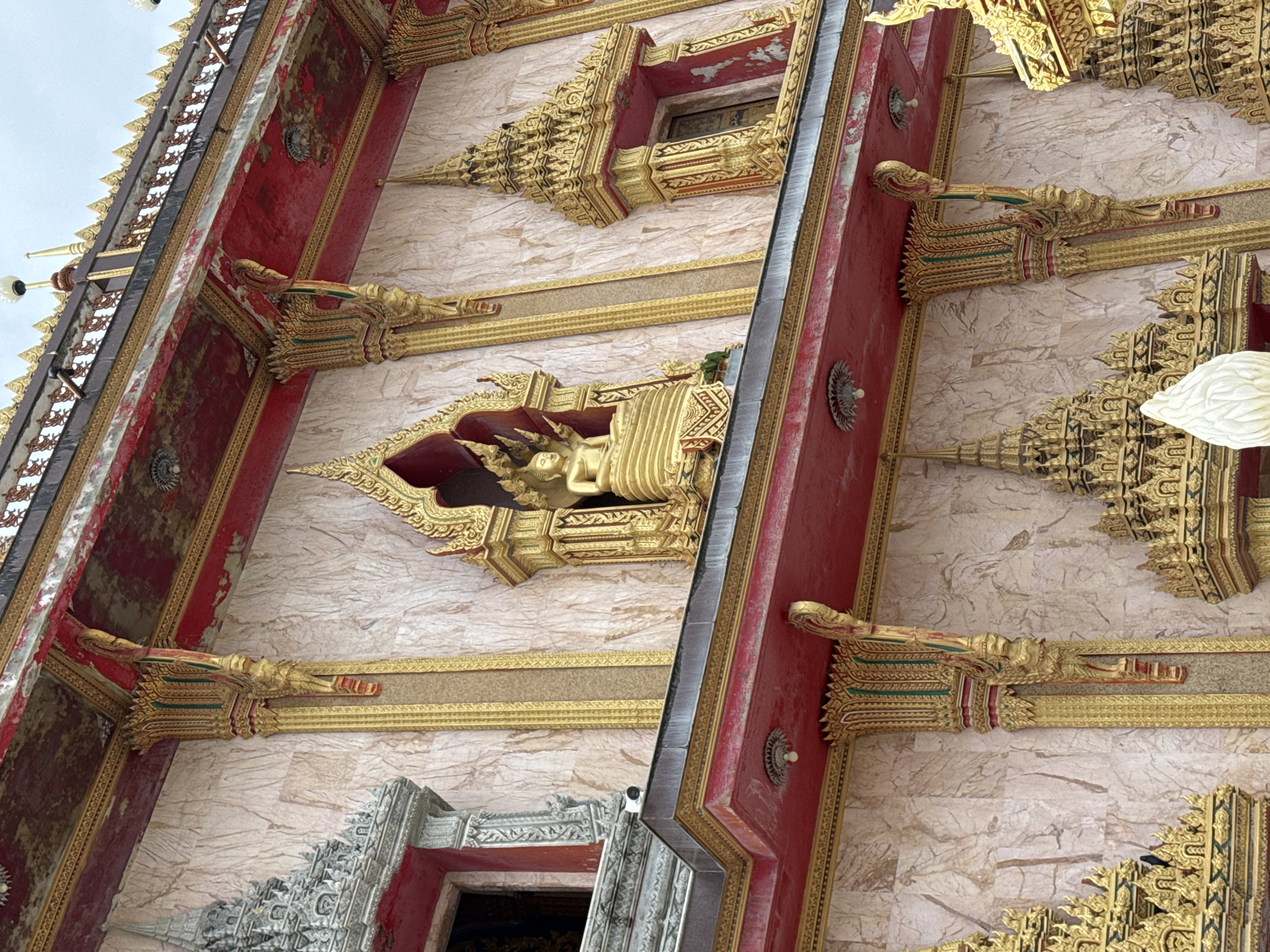 The image shows a close-up, low-angle view of the facade of a temple in Chalong, Thailand. The architecture is ornate, with intricate golden detailing against a light pink marble-like background. Red painted eaves with golden trim run horizontally across the structure. A golden statue of Buddha is visible in a niche above the eaves. Decorative golden spires and elaborate carvings adorn the building. The sky is overcast.