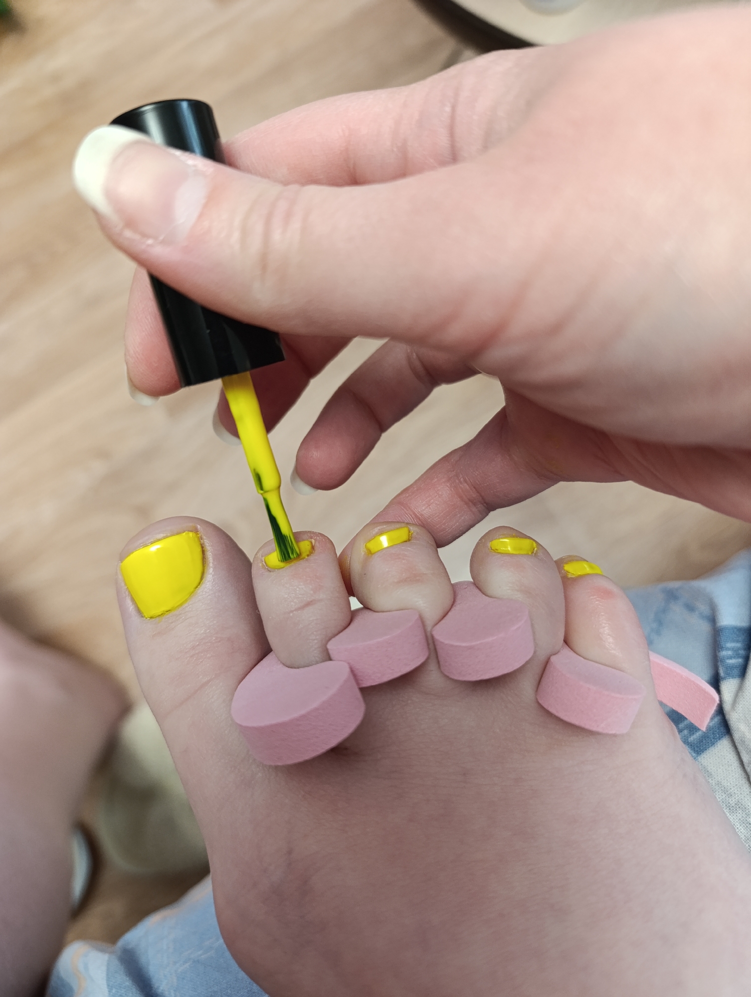 A close-up shot depicts a person painting their toenails with yellow nail polish. The foot is isolated with pink foam toe separators. A hand holds a black nail polish bottle with a yellow brush applying the polish. The large toe is fully painted, while the others have partial applications. The background is a neutral wooden floor and a glimpse of blue patterned fabric. The location is unknown.