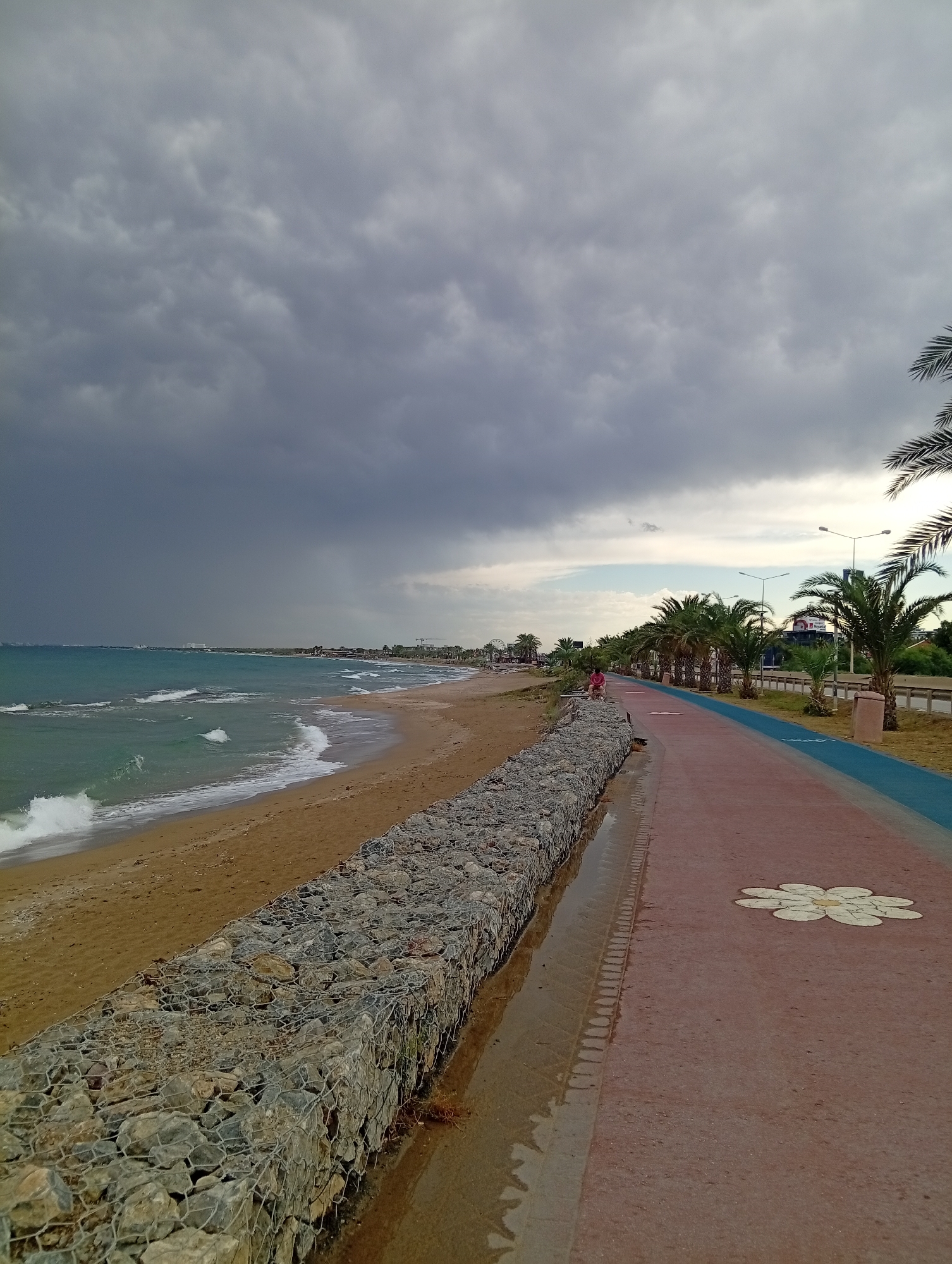 The image depicts a coastal promenade in Trikomo, Northern Cyprus. The sky is overcast with dark, heavy clouds, suggesting an impending storm or recent rainfall. The sea is a turquoise color, with moderate waves crashing onto a sandy beach. The beach is bordered by a gabion wall constructed of large stones encased in wire mesh. Adjacent to the wall is a paved promenade, with separate lanes for walking and cycling. The walking path is reddish-brown, and features a white flower mosaic design. A blue lane for cycling is next to the walking path. Palm trees line the promenade, and a road with streetlights is visible further inland. A single person, wearing a red shirt, is seated on a bench on the promenade, facing the sea. The overall atmosphere is moody and dramatic due to the weather.