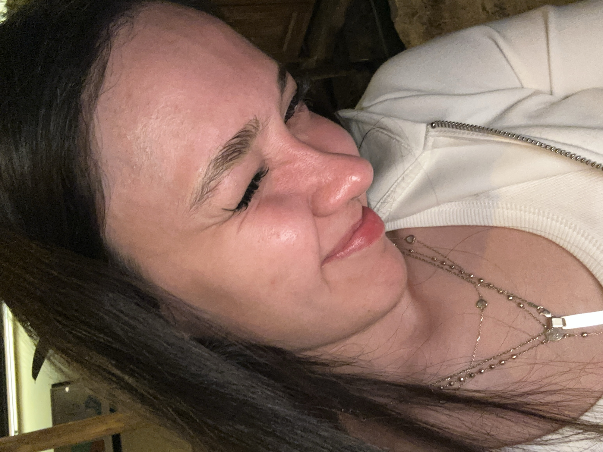 A close-up, rotated image shows a fair-skinned woman with dark hair. She's wearing a white zip-up hoodie and a layered silver necklace. Her eyes are closed, and her face exhibits a slight grimace. The background is indistinct, but hints suggest an interior setting with wooden elements. The location in Yerevan, Armenia, is unknown.