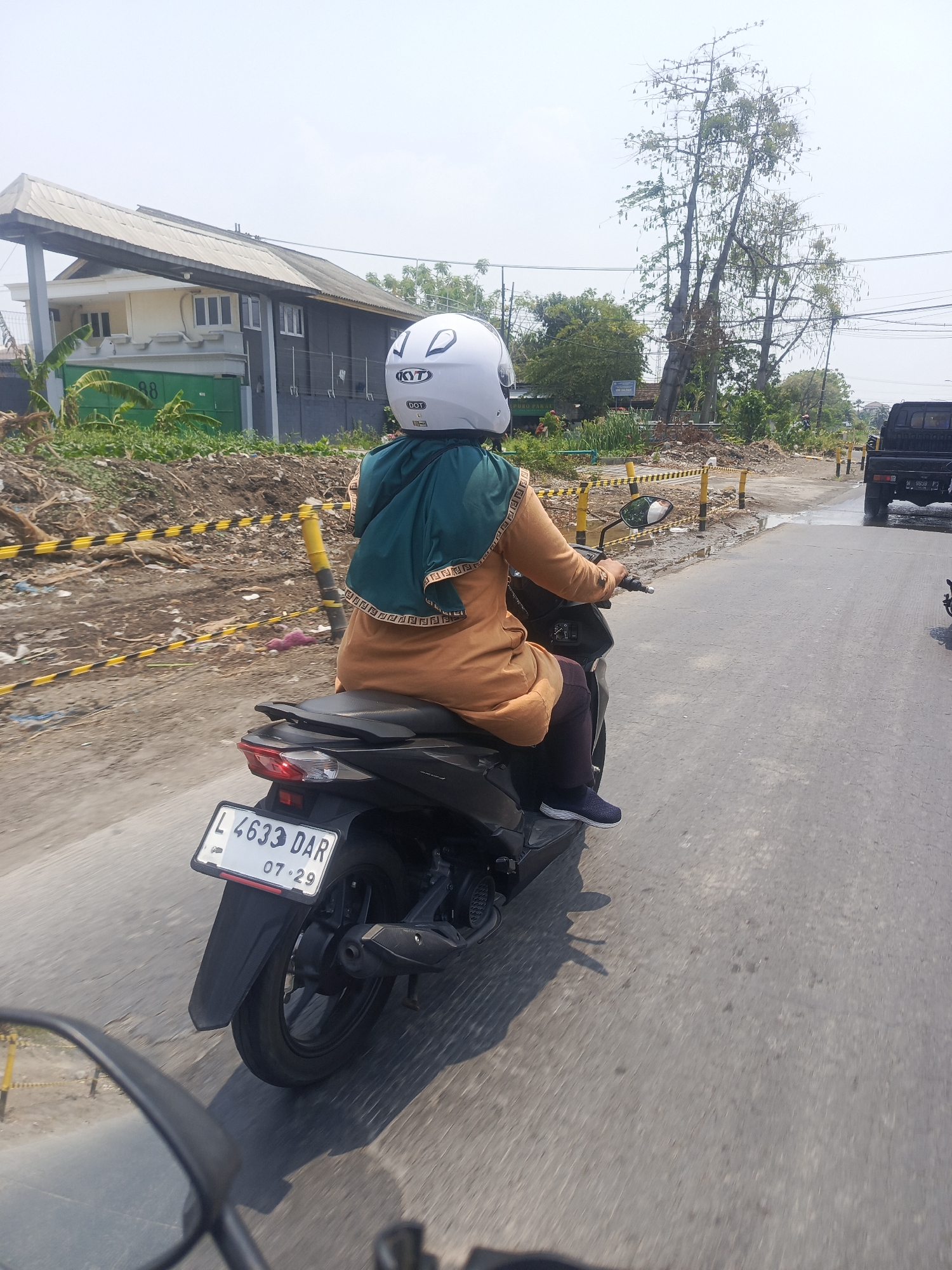 The image depicts a side view of a person riding a dark-colored motorcycle on an asphalt road. The rider is wearing a white Kyt helmet and a turquoise headscarf over an orange tunic. Their right hand is on the handlebar, and their legs are positioned to ride the motorcycle. The motorcycle's license plate reads "L 4633 DAR 07-29".
In the background, there is a building with a light-colored facade and a dark-colored upper section, along with a green gate. Yellow and black striped construction barriers are visible on the left side of the road, indicating ongoing construction or road work. Several trees, some appearing dry and tall, are present behind the barriers. A black pickup truck is visible further down the road on the right. The sky is bright and overcast, suggesting daytime.
The scene is consistent with a street in an urban or semi-urban area of Surabaya, Indonesia, given the presence of common Indonesian motorcycle license plate format and the general landscape. The road appears wet in places, and there is debris scattered along the roadside.