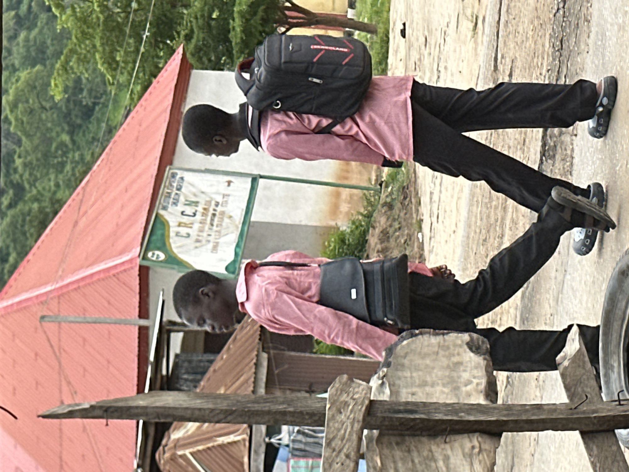 Two young boys are walking on a dirt path in front of a building with a red corrugated metal roof in Mararaba, Nigeria. The boy on the left is wearing a pink long-sleeved shirt and black pants, carrying a black messenger bag across his body. The boy on the right is also wearing a pink long-sleeved shirt and black pants, and he has a black backpack on. In the background, there is a sign with green and white text. Trees and vegetation are visible behind the building. The ground is uneven and appears to be unpaved.