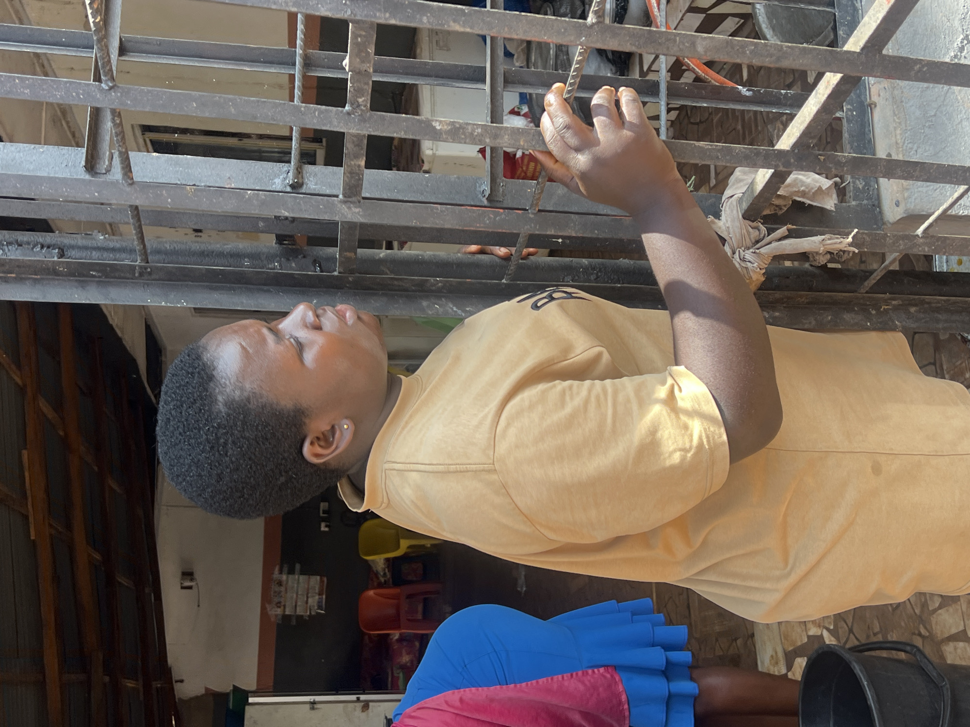 A young woman of African descent with short, dark hair and a golden earring in her left ear is standing by a metal gate, wearing a tan t-shirt with a black logo. She is looking to her right, with her right hand resting on a horizontal bar of the gate. Her left arm is bent, with her hand near her chest. Behind her, partially visible, is another woman wearing a bright blue skirt and a pink shawl. A black bucket is on the ground to the left of the frame. The background shows a building with a corrugated metal roof, wooden beams, and what appears to be a shop or open-air market with chairs and some items on display. The scene is outdoors, with natural lighting.