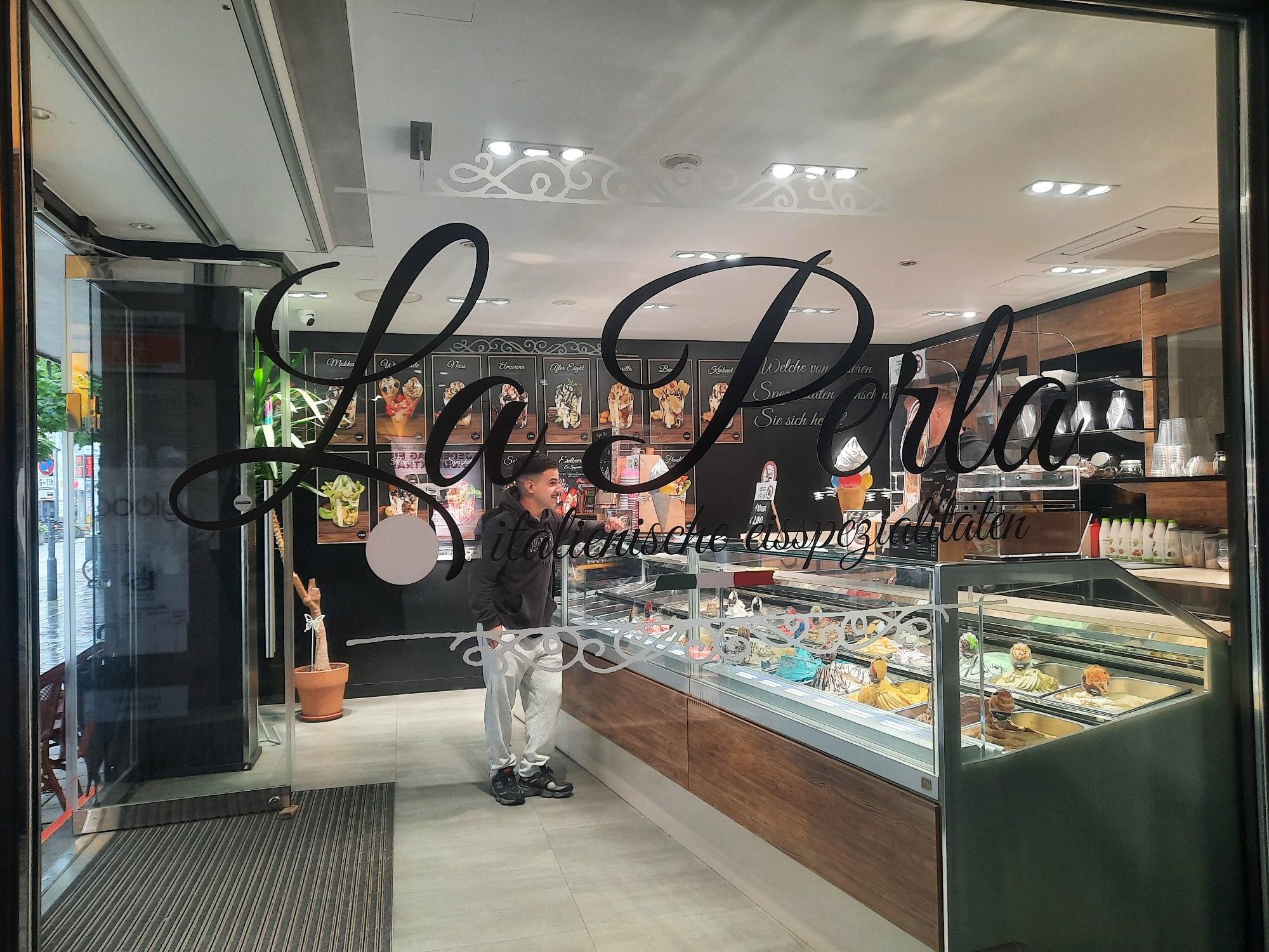 The image shows the exterior of an ice cream shop called "La Perla" located in Rosenheim, Germany, as indicated by the store's signage. The shop's name is prominently displayed in large, stylized black lettering across the glass facade. Through the glass, an ice cream display case is visible, filled with various flavors of gelato. A young man, dressed in a grey hoodie and light-colored track pants, stands inside the shop, looking towards the display case and appearing to interact with something out of frame to the right. The shop's interior features black walls adorned with pictures of ice cream creations and some German text, likely menu items or slogans. The lighting inside appears bright, and the overall atmosphere suggests a typical ice cream parlor. The exterior shows a glass door and a mat on the ground, typical of a shop entrance.