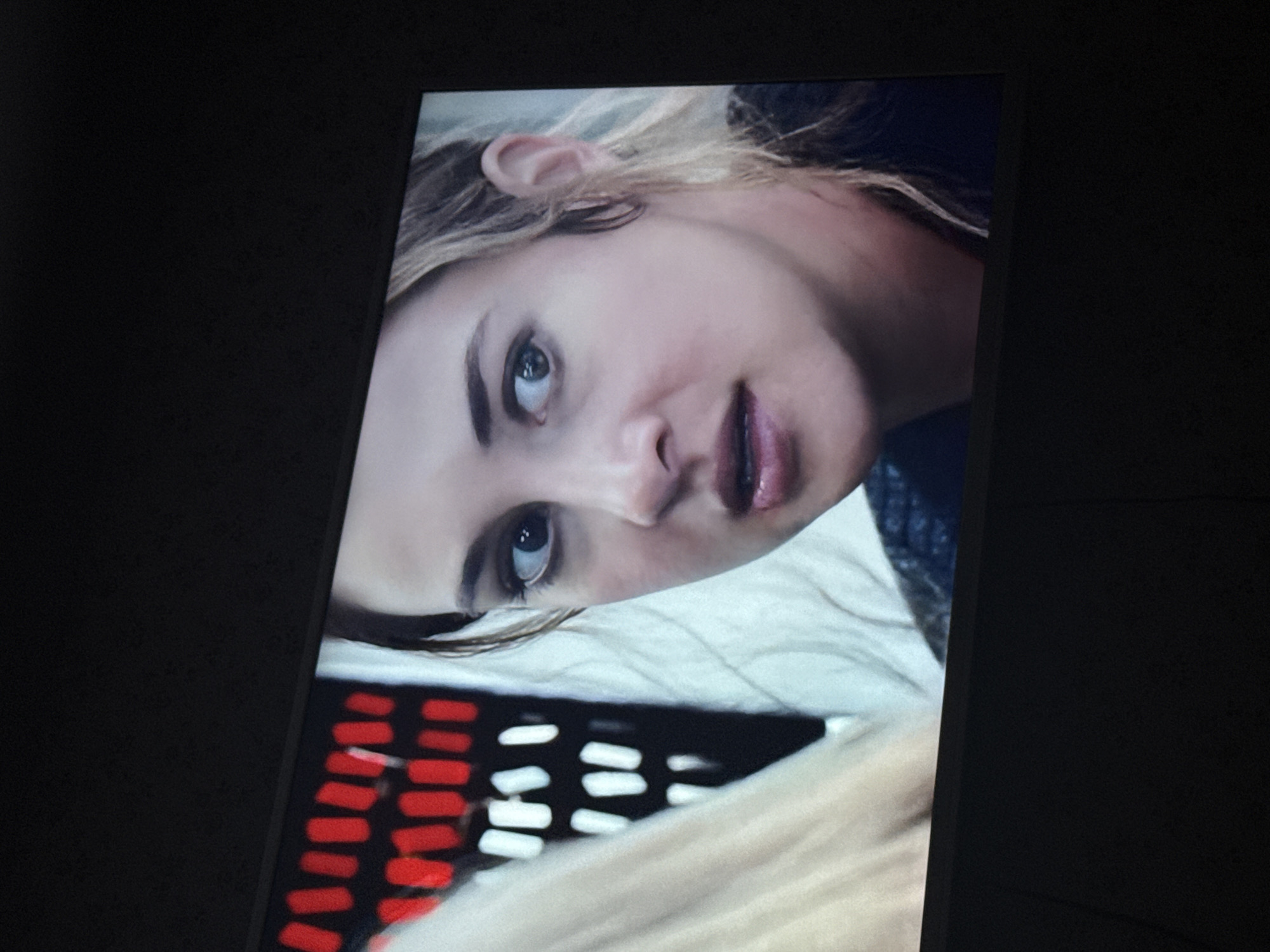 A close-up shot of a TV screen displays a blonde woman with her head tilted, her eyes gazing towards the upper left of the screen. Her hair is disheveled, and she wears a dark shirt. The background includes a white, textured surface, possibly a pillow, and a blurred pattern of red and white shapes, likely part of a design or image on the screen. The TV screen is surrounded by dark surroundings. The location in the city of Mytishchi, Russia is not possible to be determined from the picture.