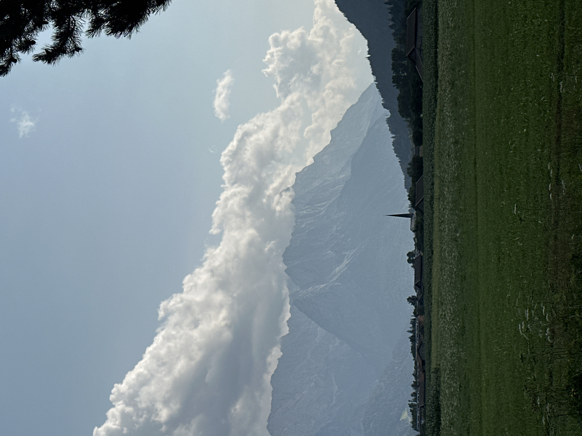 The image shows a wide shot of a rural landscape with mountains in the background and a field in the foreground. The mountains are layered and hazy, with large white clouds partially obscuring them. In the middle ground, there is a village with houses and a church steeple visible. A dark green, lush field occupies the foreground, with some wild flowers and vegetation. The sky is a light blue with scattered clouds. The scene appears to be a view of Farchant, Germany, with the Alps in the background.
