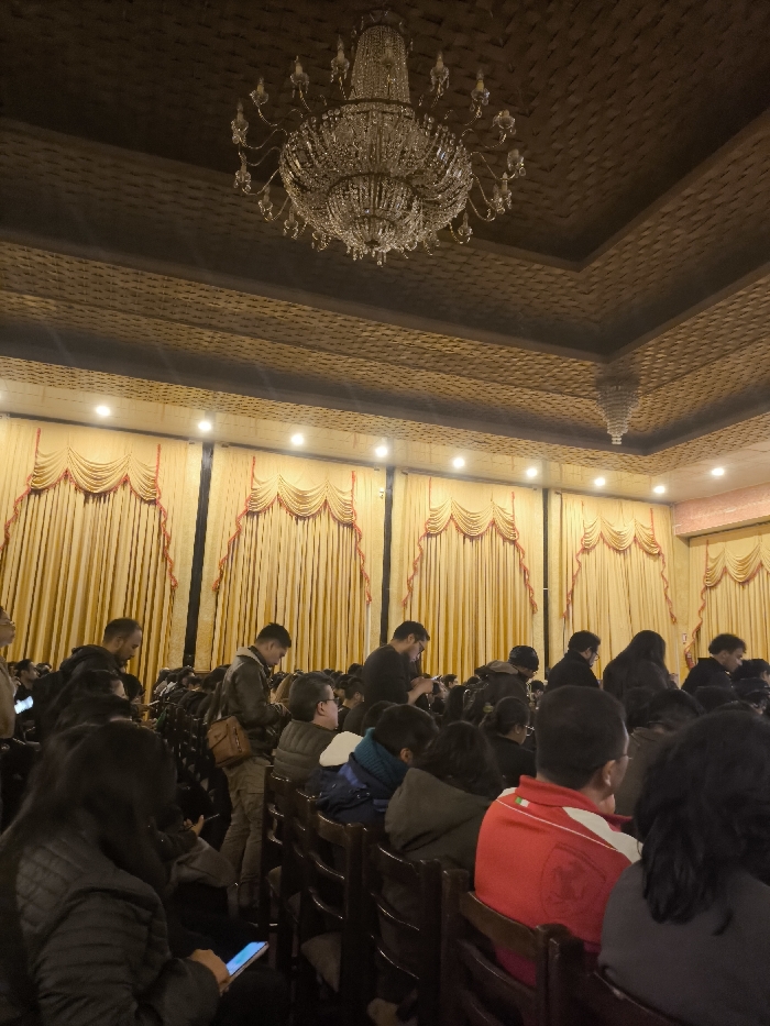 The image depicts an indoor scene within a large, formal hall or auditorium in La Paz, Bolivia.
**Environment:** The upper third of the frame is dominated by a dark, intricately patterned ceiling, possibly coffered or carved wood, from which a large, multi-tiered crystal chandelier hangs centrally. Recessed white spotlights are visible along the ceiling's perimeter. A reflection of a second, smaller chandelier or light fixture appears in the upper right. The walls are covered with floor-to-ceiling golden-yellow drapes, featuring scalloped valances trimmed with red piping and vertical red ties that create a decorative pattern. Additional recessed spotlights illuminate the drapes from above. The flooring is not visible.
**People:** A seated audience occupies the foreground and midground, visible from the back and sides. Approximately 20-30 individuals are discernible. They are predominantly adults, dressed in casual to semi-casual dark attire, including jackets and sweaters. Most are seated in rows of dark wooden chairs, facing forward, indicative of an audience attending an event. Several individuals, particularly in the foreground and midground, are actively using illuminated smartphones, looking down at their screens. A few people are standing in the aisles or at the rear of the seating area.
**Objects:** Key objects include the prominent crystal chandelier, the rows of dark wooden chairs, and numerous illuminated smartphones held by attendees. One individual has a light brown shoulder bag.
**Actions/Interactions:** The primary actions observed are individuals seated as an audience, with many engaged with their personal electronic devices. There are no overt interactions between attendees visible.
**Location in La Paz, Bolivia:** The ornate design of the ceiling, the large crystal chandelier, and the formal drapes are characteristic of a grand public gathering space. This style suggests a location such as an auditorium, a theater hall (e.g., similar to spaces found within the Teatro Municipal Alberto Saavedra Pérez), a large conference room, or a ballroom within a prominent institutional building or hotel in La Paz.
