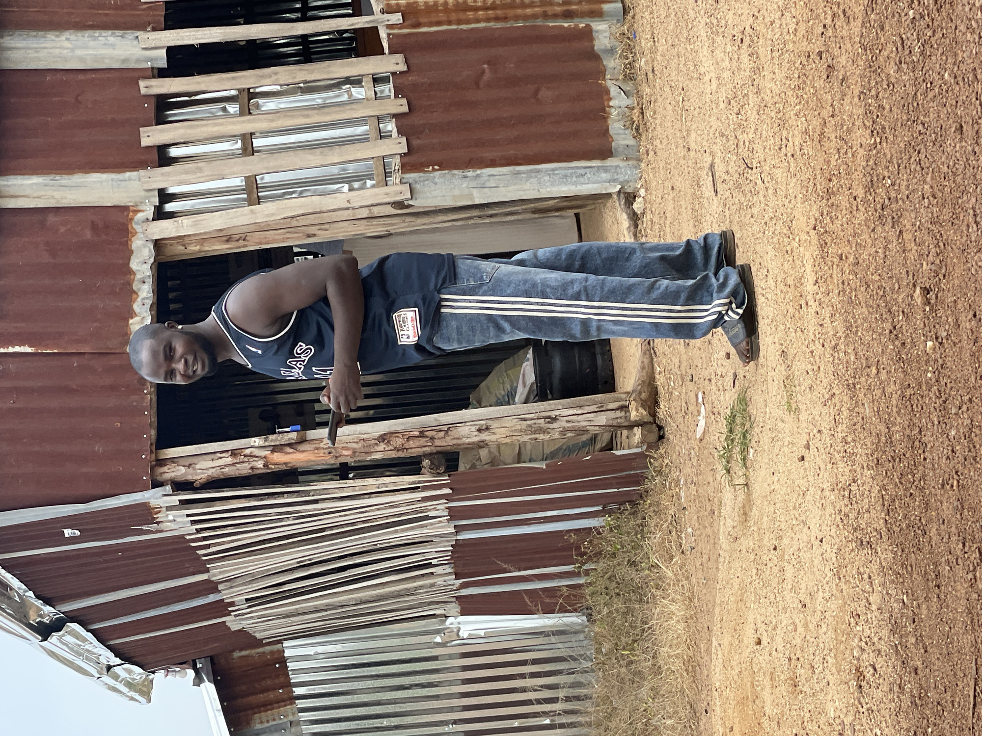 A young Black man stands in front of a rustic, partially constructed building. The building appears to be made of corrugated metal and unfinished wood, with some sections appearing weathered and rusty. The man is wearing a dark blue tank top with white stripes on the straps and "LAS" visible in white letters. He is also wearing blue jeans with white stripes down the sides and dark sandals. He is holding a small, dark object in his right hand. The ground is covered in sandy dirt and dry grass. The lighting suggests it is daytime, and the overall impression is of a rural or undeveloped setting. The man's expression is neutral.