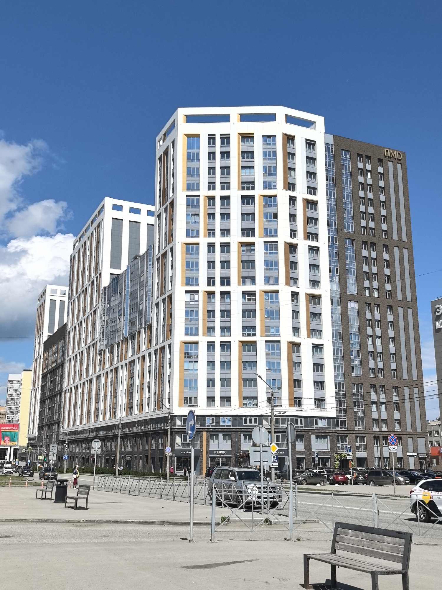 The image shows a cityscape in Perm, Russia, under a bright blue sky with scattered white clouds. Prominently featured are modern apartment buildings with a mix of white, brown, and grey facades, and numerous windows. One of the taller buildings has the letters "ПМД" (PMD) visible at the top.
In the foreground, there's a paved area with metal fencing and wooden benches. A few people are visible in the middle distance walking along the sidewalk. Cars are parked along the street adjacent to the buildings, and some traffic is visible. Street signs and streetlights are scattered throughout the scene. Overall, the setting appears to be a commercial or residential area within the city.