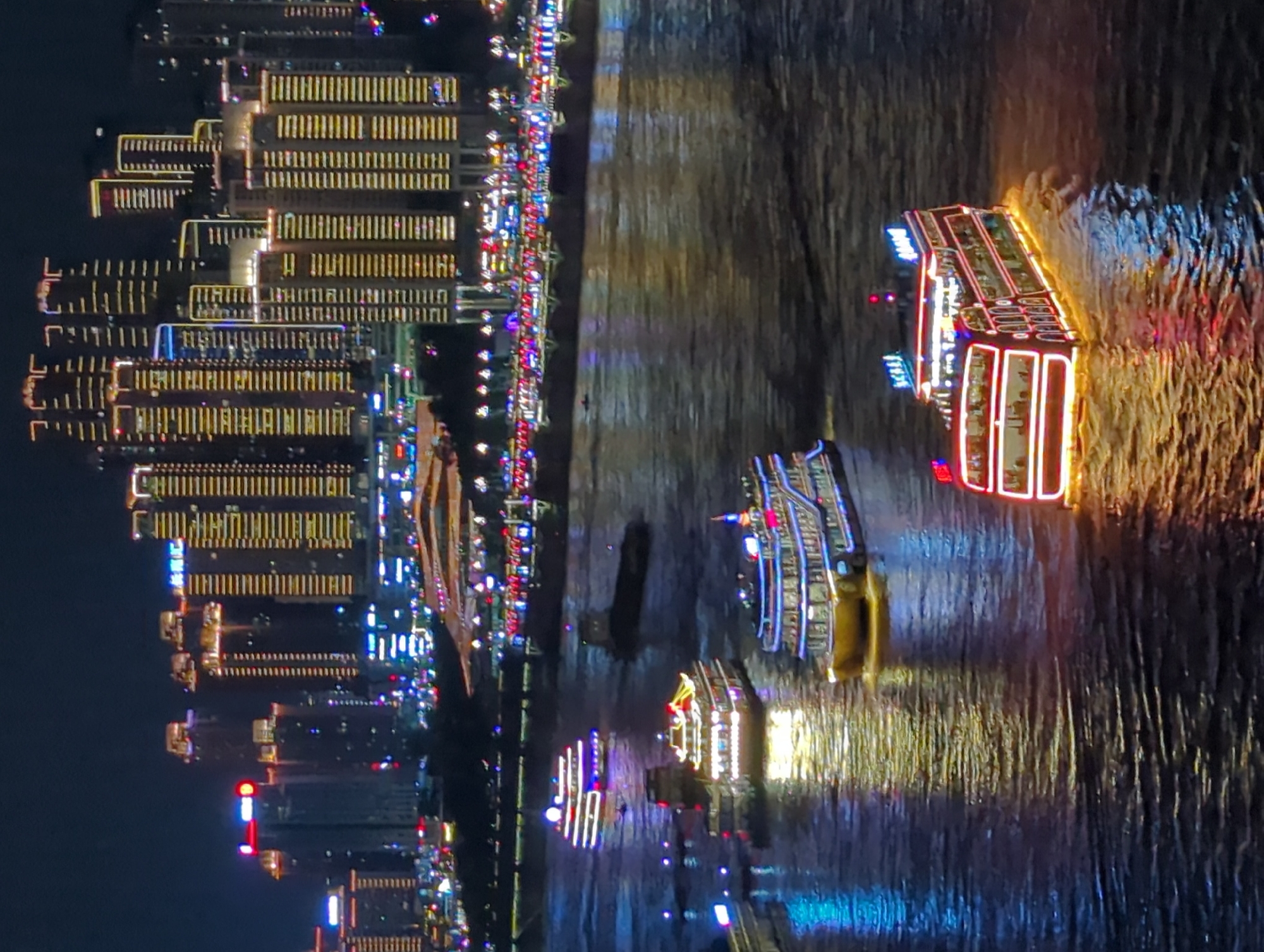 The image depicts a nighttime view of the Yuzhong District in China, specifically focusing on the illuminated skyline and a body of water with several boats. Tall, modern buildings dominate the background, adorned with numerous bright lights that outline their structures and windows, creating a vibrant urban panorama. The water in the foreground is reflective, mirroring the city lights and the illuminated boats.
Several passenger boats are visible on the water. One prominent boat in the foreground is multi-decked and extensively lit with red and white lights along its exterior and upper levels, with some blue lights also present. Another boat further back, also multi-decked, is illuminated with yellow and white lights, displaying a traditional architectural style on its roof. A third, smaller boat, is lit with red, white, and blue lights. In the distance, other vessels and the reflections of the cityscape suggest active waterway traffic. The overall scene conveys a lively and illuminated urban environment at night.